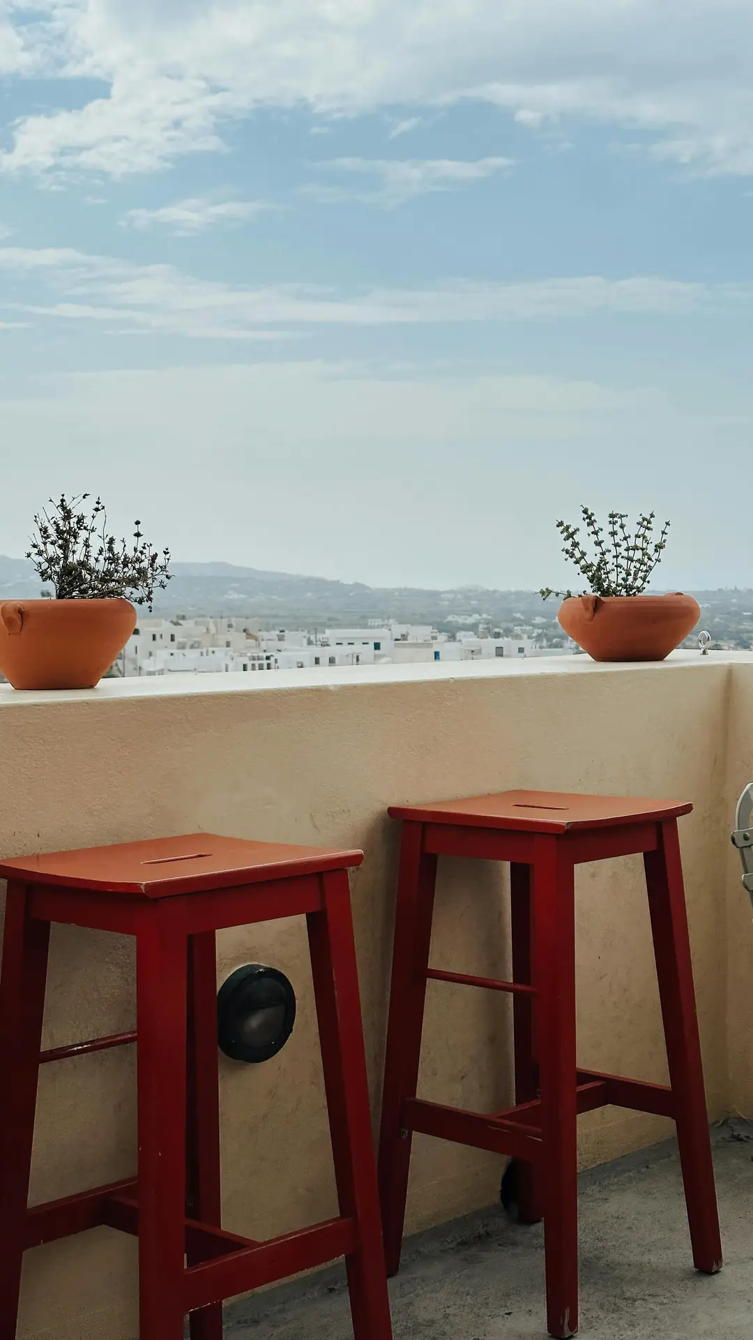 A minimal balcony setup with two bright red stools placed against a cream railing, decorated with terracotta pots holding small green plants. Overlooking a townscape with hills in the distance, this scene highlights simple yet impactful balcony decoration ideas for adding color and charm to compact spaces.