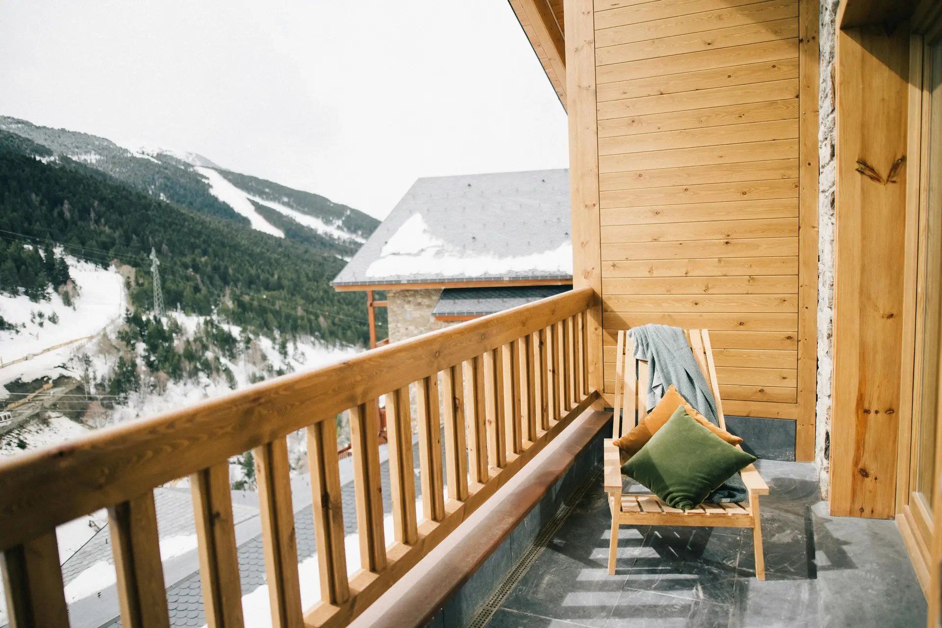 A wooden balcony overlooking snowy mountain slopes, styled with a simple wooden lounge chair topped with soft pillows and a blanket, perfect for sunbathing in winter. A cozy example of balcony decoration ideas that balance warmth and comfort with stunning alpine views.
