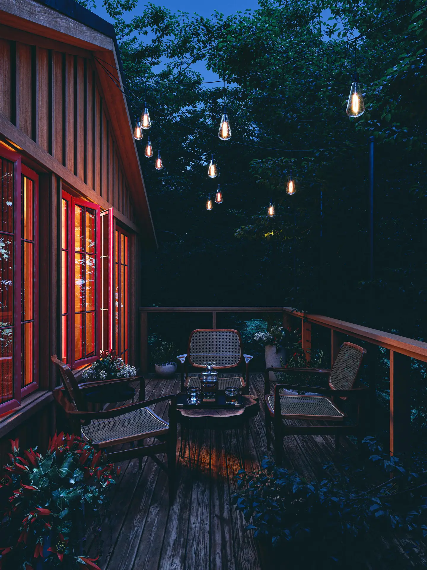 A cozy evening balcony scene showcasing three seats with wooden table and fairylights, surrounded by lush greenery. Warm glowing string lights hang above, casting a soft atmosphere, while the rustic wood deck and red-framed windows add charm. Perfect for inspiring balcony decoration ideas that combine comfort with ambient lighting for intimate outdoor gatherings.