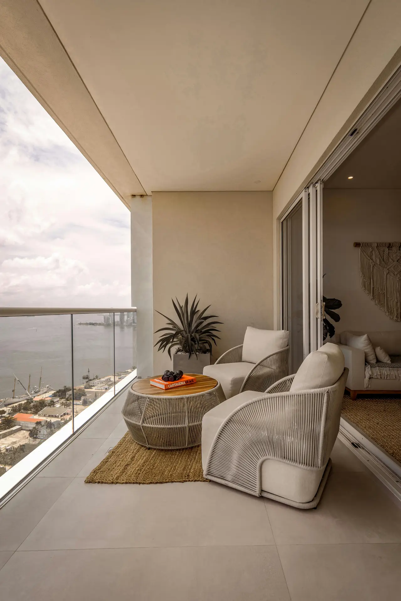 Modern balcony overlooking the ocean, furnished with two big chairs with geometric table set on a natural woven rug. The clean lines, neutral tones, and glass railing create a sleek atmosphere, offering stylish balcony decoration ideas for anyone seeking a mix of comfort and contemporary design.