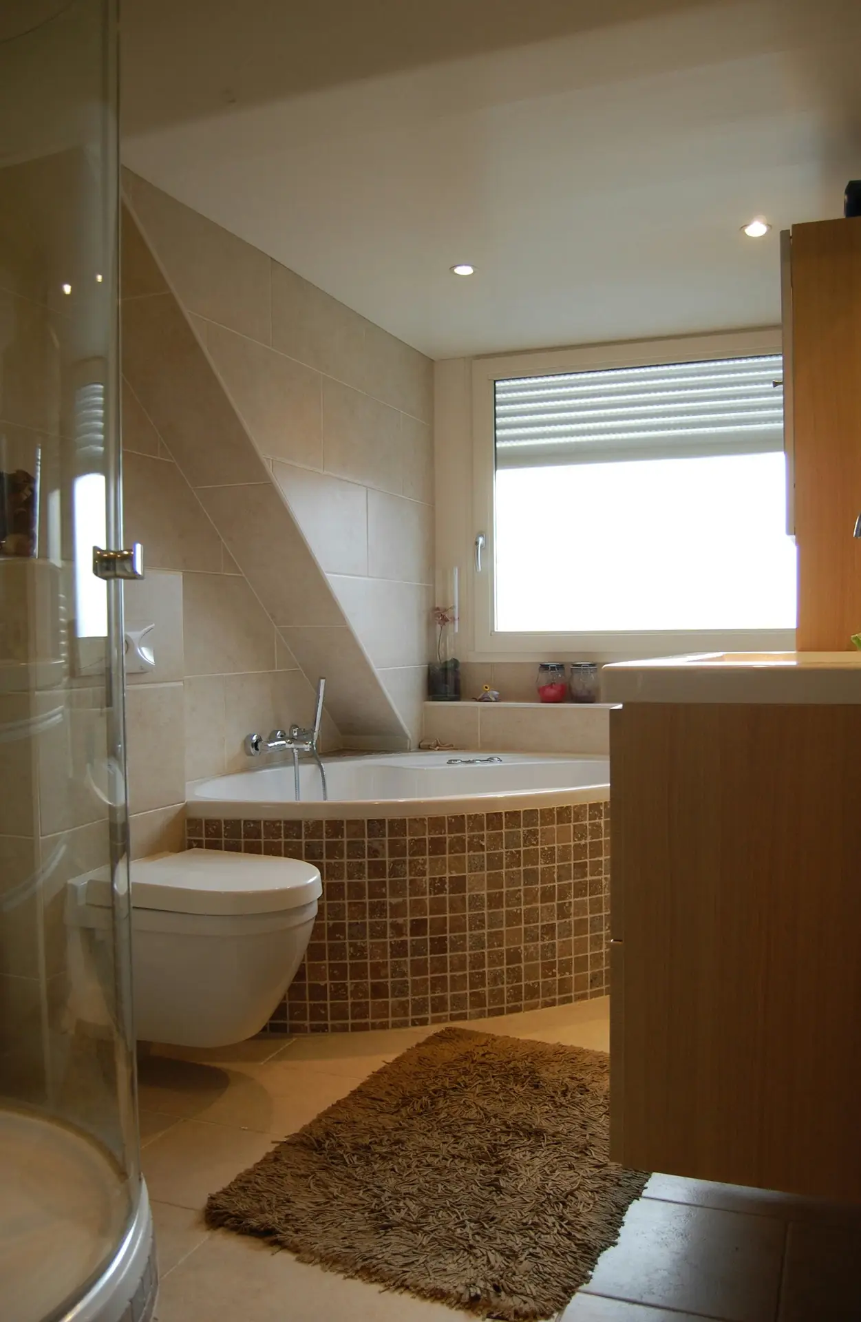 Modern bathroom featuring beige wall tiles with brown mosaic bathtub surround, a wall-mounted toilet, and a corner bathtub beneath a slanted ceiling, offering bathroom tiles ideas that combine warm tones with smart space utilization.