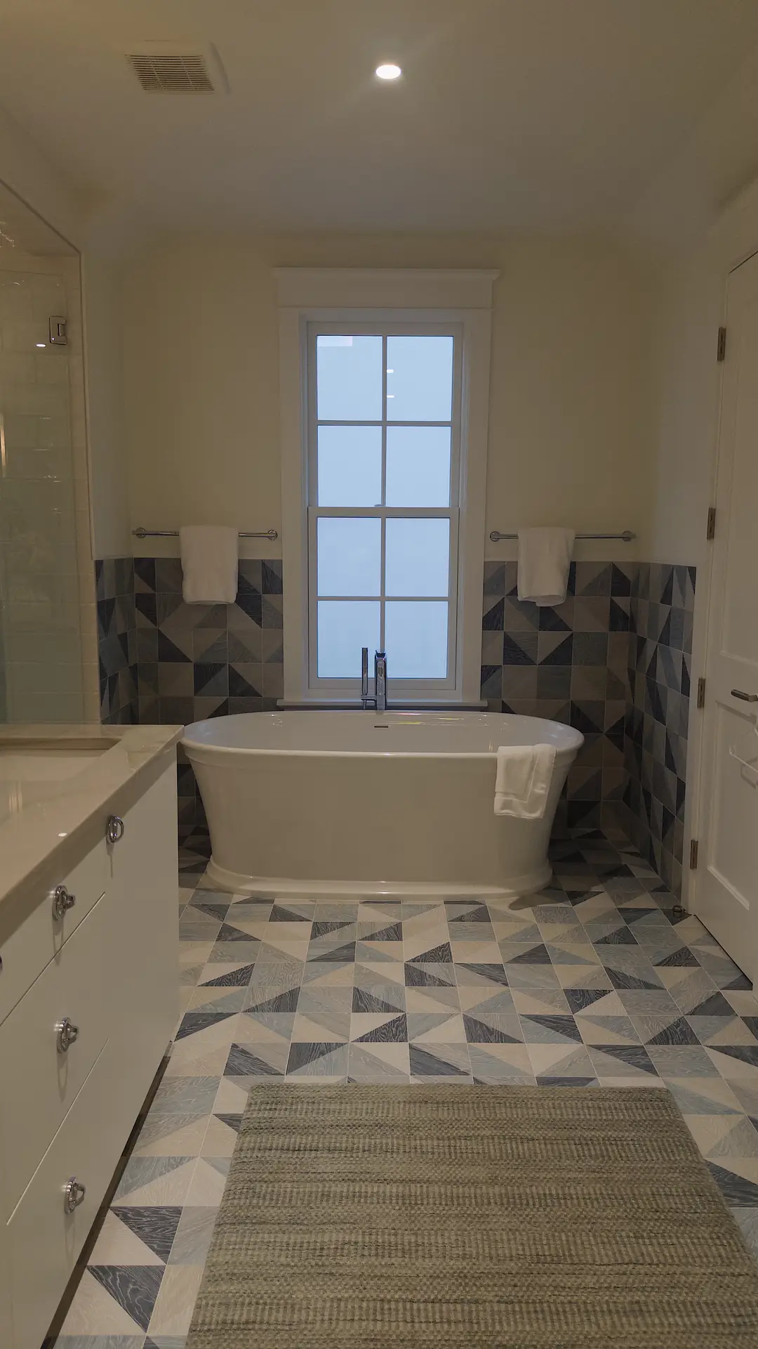 Modern bathroom featuring bathroom tiles ideas with Geometric Patterned Tiles In Blue & Gray Tones that flow seamlessly from the floor up the lower half of the walls. The bold triangular design adds movement and character, complementing the sleek freestanding bathtub centered under a bright window. A light vanity with polished handles and a simple neutral rug balance the striking tilework, making this space both contemporary and inviting.