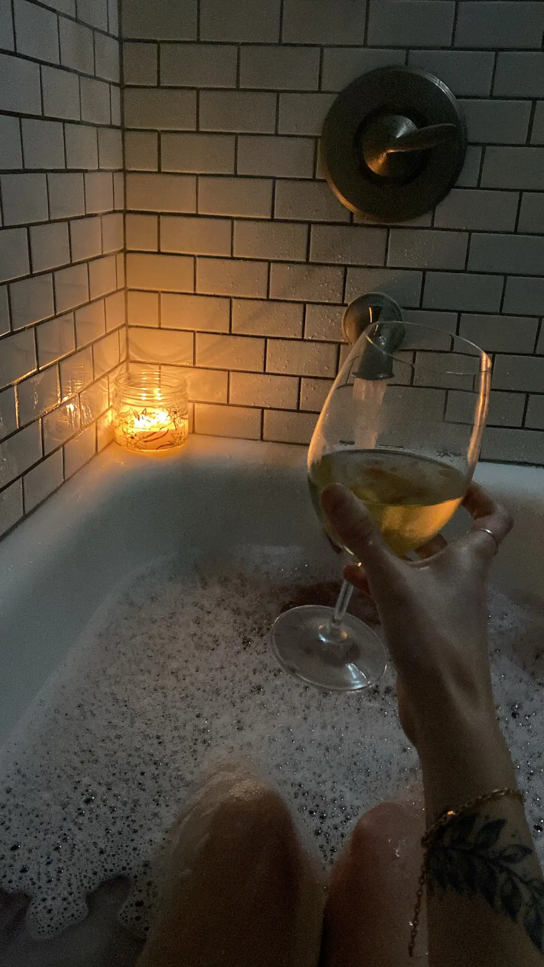 Cozy bathtub moment with bubbles, a lit candle, and a glass of wine, set against glossy white subway tiles that reflect the warm glow of the light-an inviting scene that doubles as inspiration for bathroom tiles ideas combining timeless design with a relaxing atmosphere.