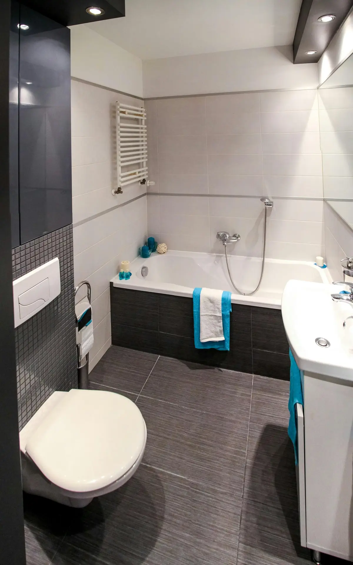 A sleek modern bathroom showcasing bathroom tiles ideas with gray large floor tiles with white rectangular wall tiles. The bathtub is clad in dark tiles that contrast against the crisp white walls, while turquoise towels and decor add a fresh accent. Recessed lighting, a wall-mounted toilet, and a compact vanity make the space both stylish and functional.
