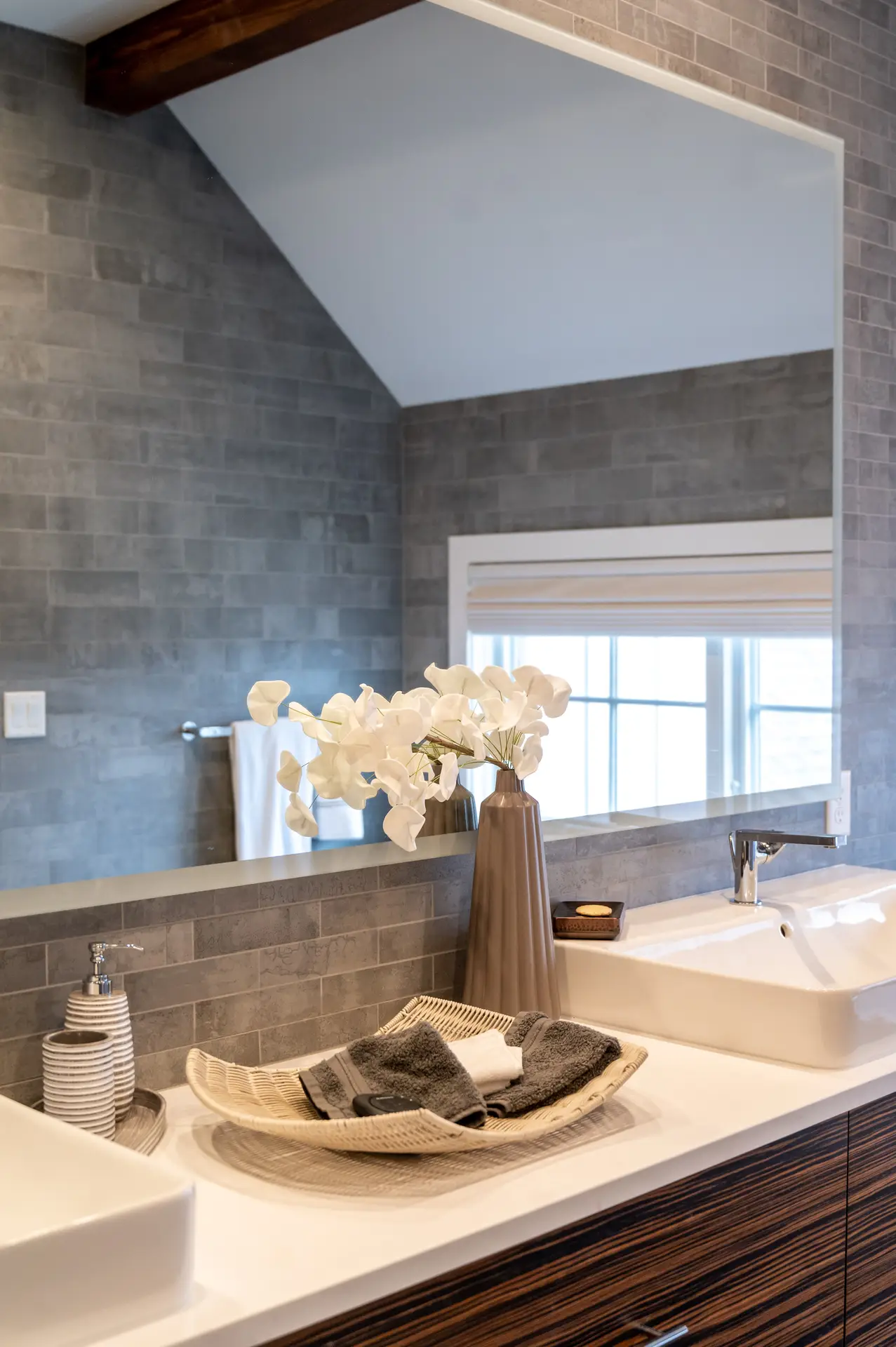 Stylish vanity setup featuring bathroom tiles ideas with Matte Gray Subway Tiles With Stone Texture, creating a natural and modern backdrop. The wall tiles extend up to the sloped ceiling, paired with a large mirror, a sleek white basin, and decorative accents like a tall vase with white flowers and a woven tray holding towels. This design blends warmth and minimalism, making it a versatile inspiration for contemporary bathrooms.