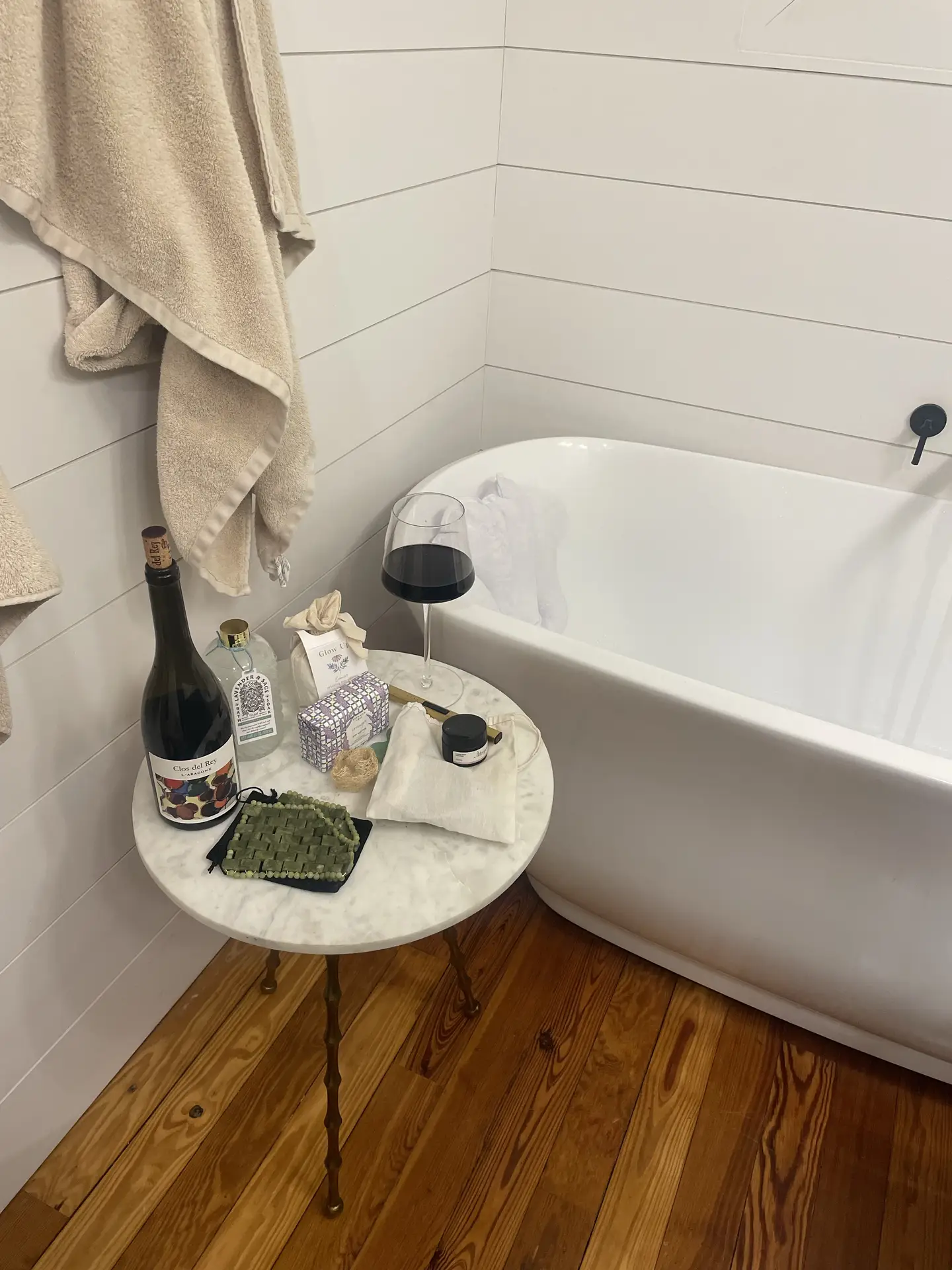 Cozy bathroom corner with a freestanding tub against shiplap wall panels, paired with warm wooden flooring and a marble side table holding wine, candles, and bath essentials-an inspiring mix of relaxation and bathroom tiles ideas that combine rustic charm with modern comfort.