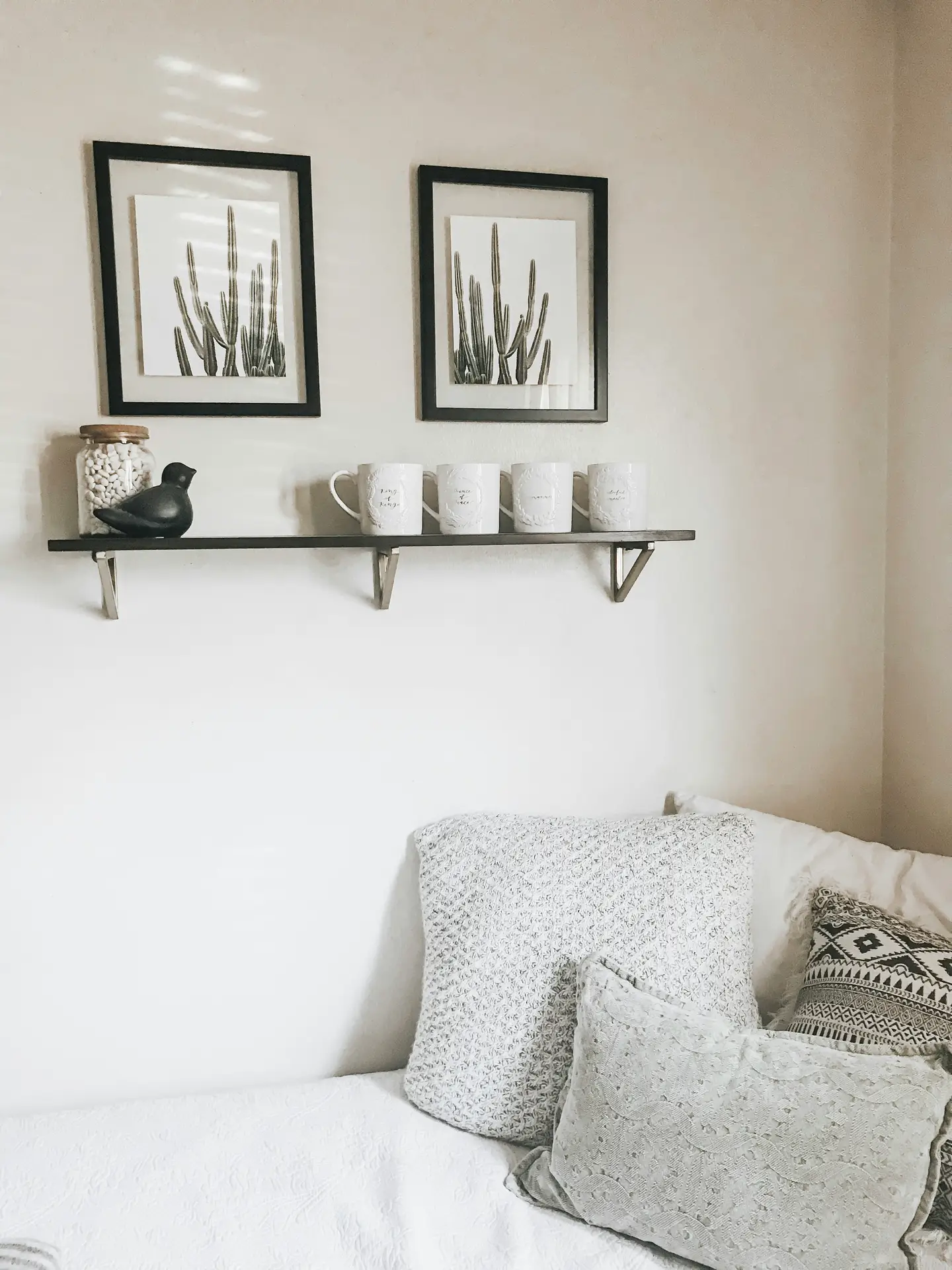 Minimalist bed decor with pillows in soft gray and neutral tones, styled in an asymmetrical arrangement that feels casual yet inviting. The mix of textured and patterned cushions adds subtle variety, while the clean white bedding keeps the look fresh. Above the bed, framed cactus prints and a simple floating shelf with mugs and small accents bring a touch of personality to the space.