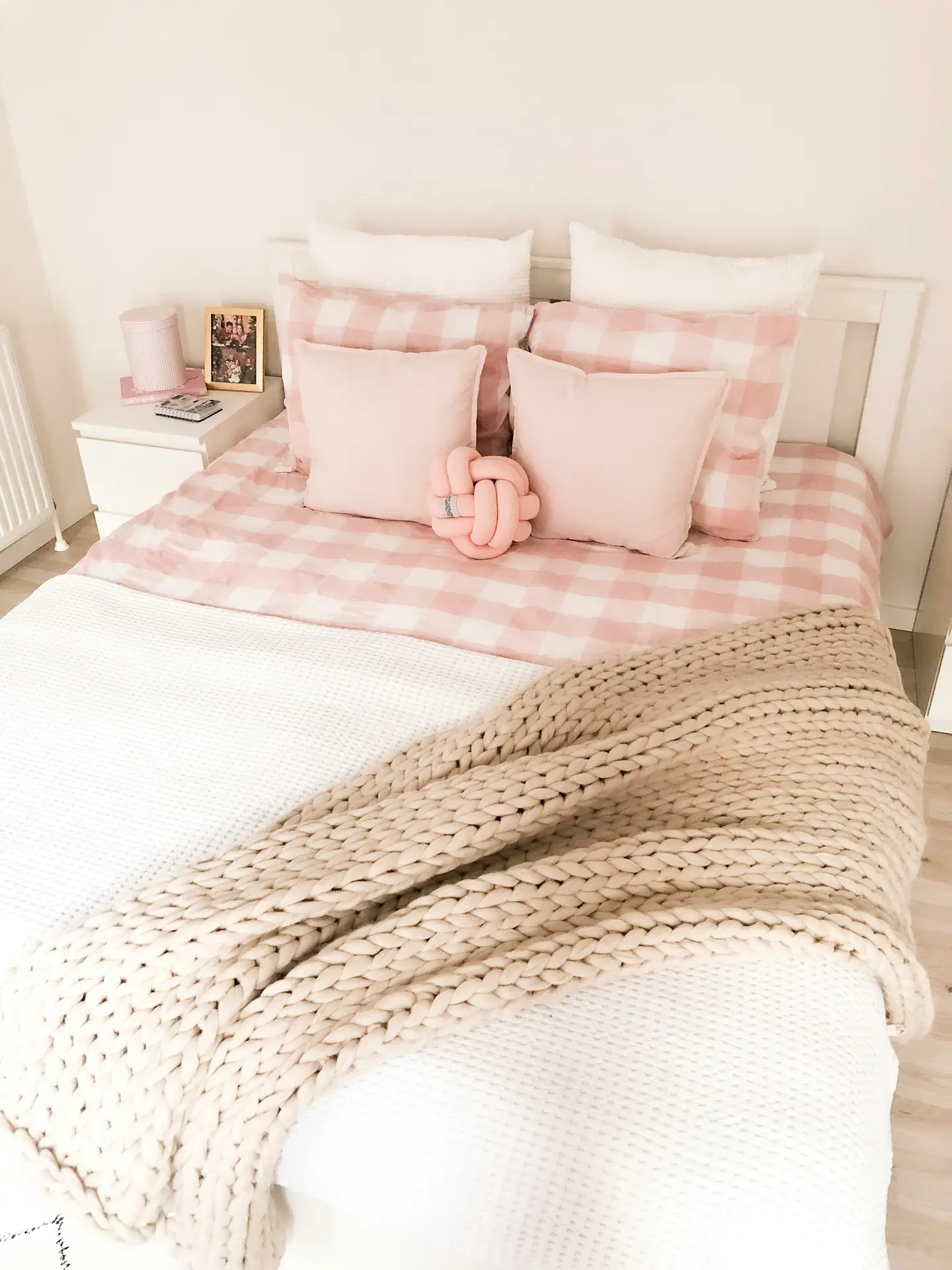 Charming bed decor with pillows styled in soft pastel tones, where blush pink cushions are paired with gingham-patterned bedding. The arrangement demonstrates a thoughtful balance between print and plain, creating harmony without overwhelming the space. A chunky knit throw draped across the bed adds cozy texture, while the small knot pillow adds a playful, modern twist.