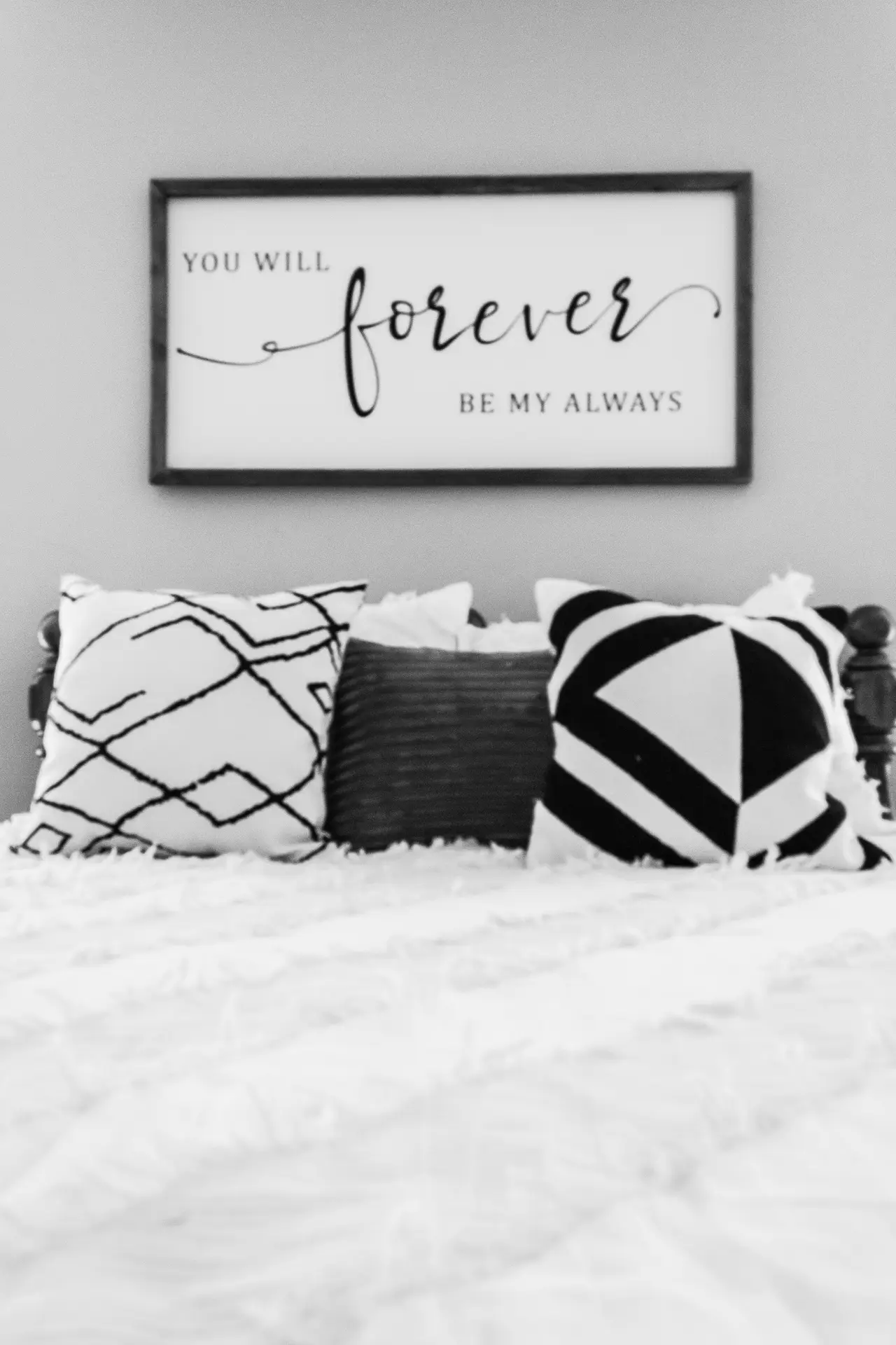 Modern bed decor with pillows styled in a monochrome palette, featuring bold black and white geometric and abstract patterned cushions. The mix of textures adds depth to the clean bedding, while the wall art above the bed with a romantic quote softens the overall look. The combination strikes a versatile balance between contemporary style and cozy charm.