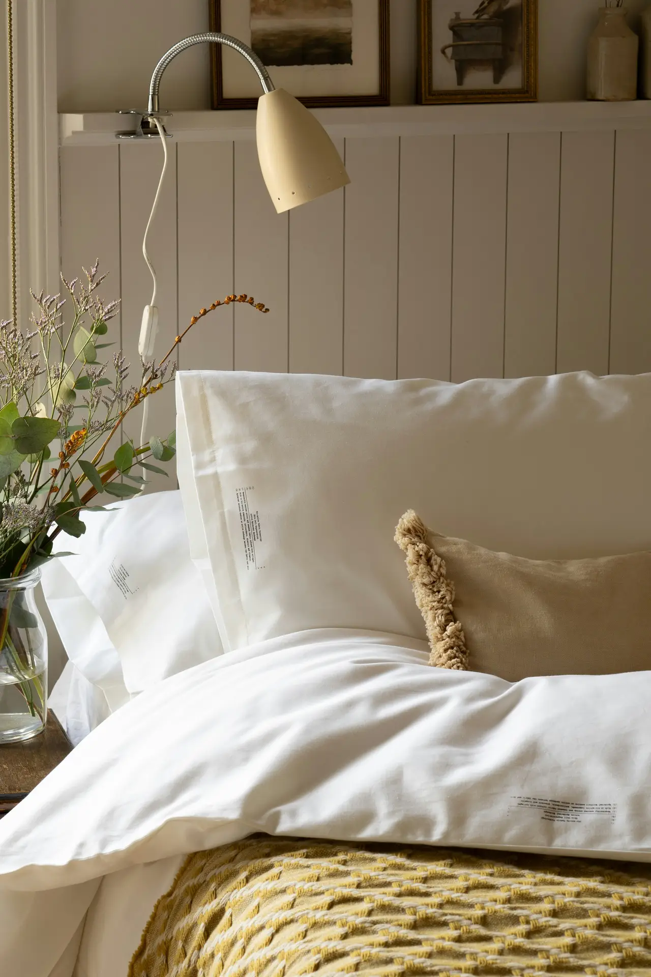 Elegant bed decor with pillows styled in soft white bedding, complemented by a beige cushion with textured trim for added charm. A mustard-yellow throw with a woven pattern introduces warmth and eye-catching details, while fresh greenery in a glass vase enhances the natural feel. A wall-mounted lamp and framed art above the headboard complete the cozy yet refined setting.