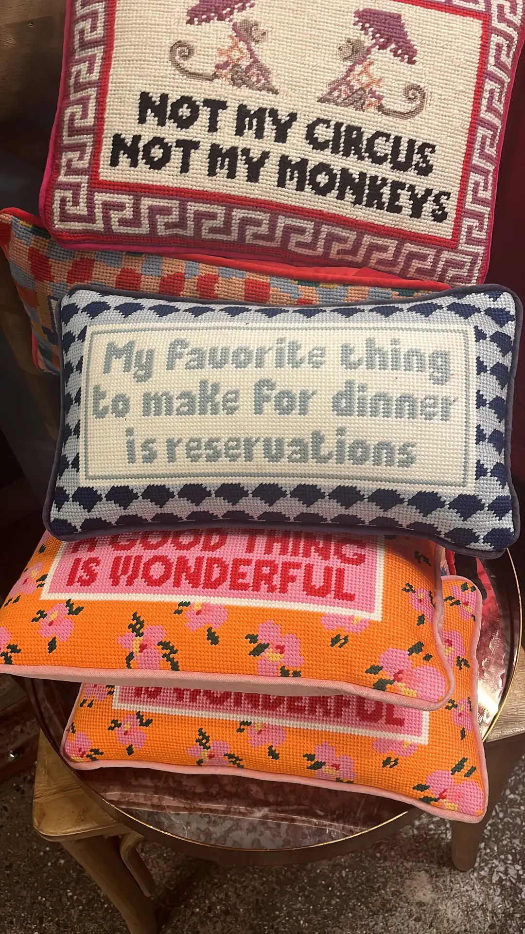 Eclectic bed decor with pillows showcasing bold needlepoint designs in bright colors and playful fonts. Each cushion is decorated with funny sayings, such as “Not my circus, not my monkeys” and “My favorite thing to make for dinner is reservations,” adding humor and personality to the setup. The mix of patterns and witty text creates a lively and versatile accent that works in both cozy bedrooms and casual living spaces.