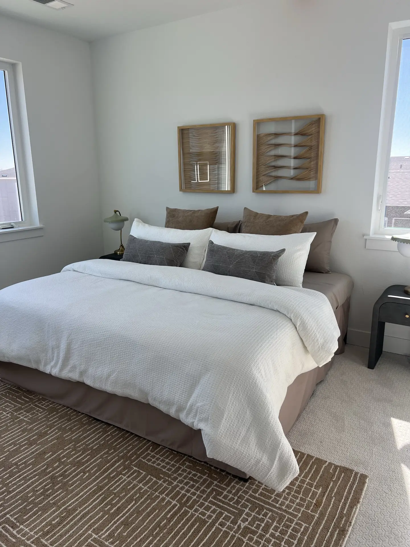 Elegant bed decor with pillows arranged in layered tones of white, gray, and brown, creating a refined balance of comfort and style. The crisp bedding and coordinated cushions evoke a luxury hotel feeling, making the room feel both inviting and sophisticated. Modern wall art, large windows, and a textured rug enhance the polished yet versatile atmosphere.
