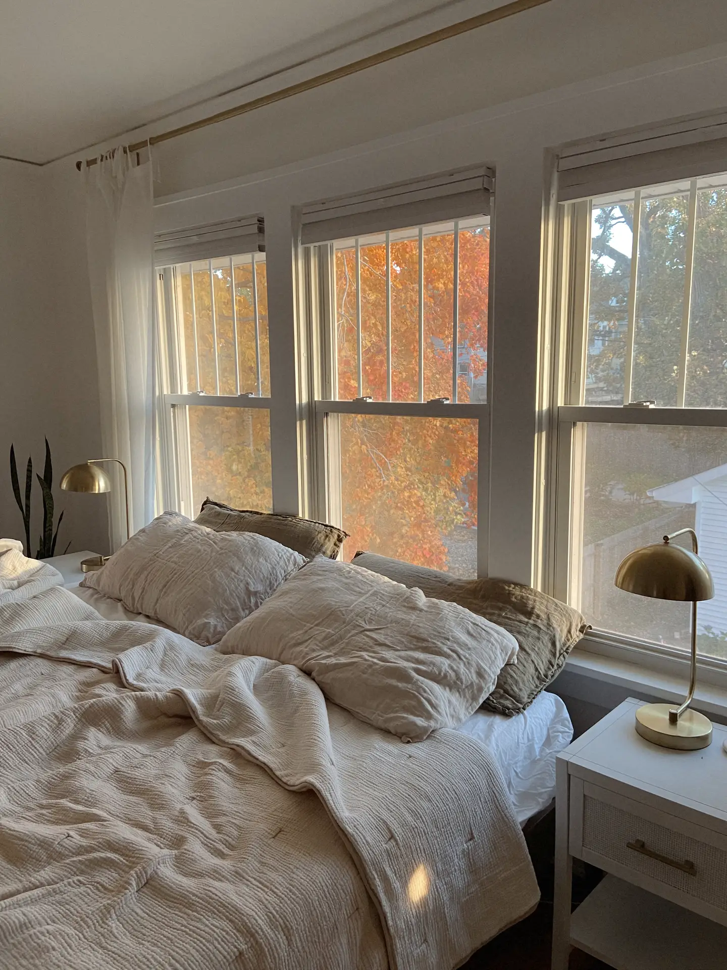 A cozy bedroom styled with layered bed decor with pillows, featuring oversized cushions in earthy tones. The bedding and soft quilt are in matching colors of beige and muted brown, creating a harmonious look. Golden lamps on white nightstands and autumn foliage visible through the large windows complete the warm, inviting atmosphere.