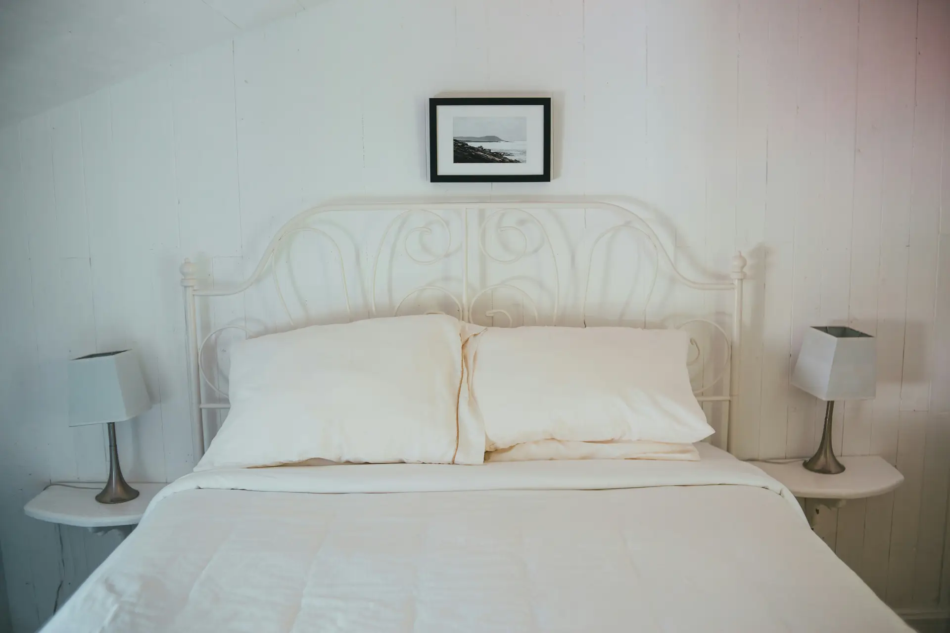 Simple bed decor with pillows styled in soft cream tones, creating a serene and calming space. The clean lines of the metal headboard, paired with matching lamps on either side, emphasize a minimalist look that feels uncluttered and airy. A single framed artwork above the bed completes the understated yet balanced design.