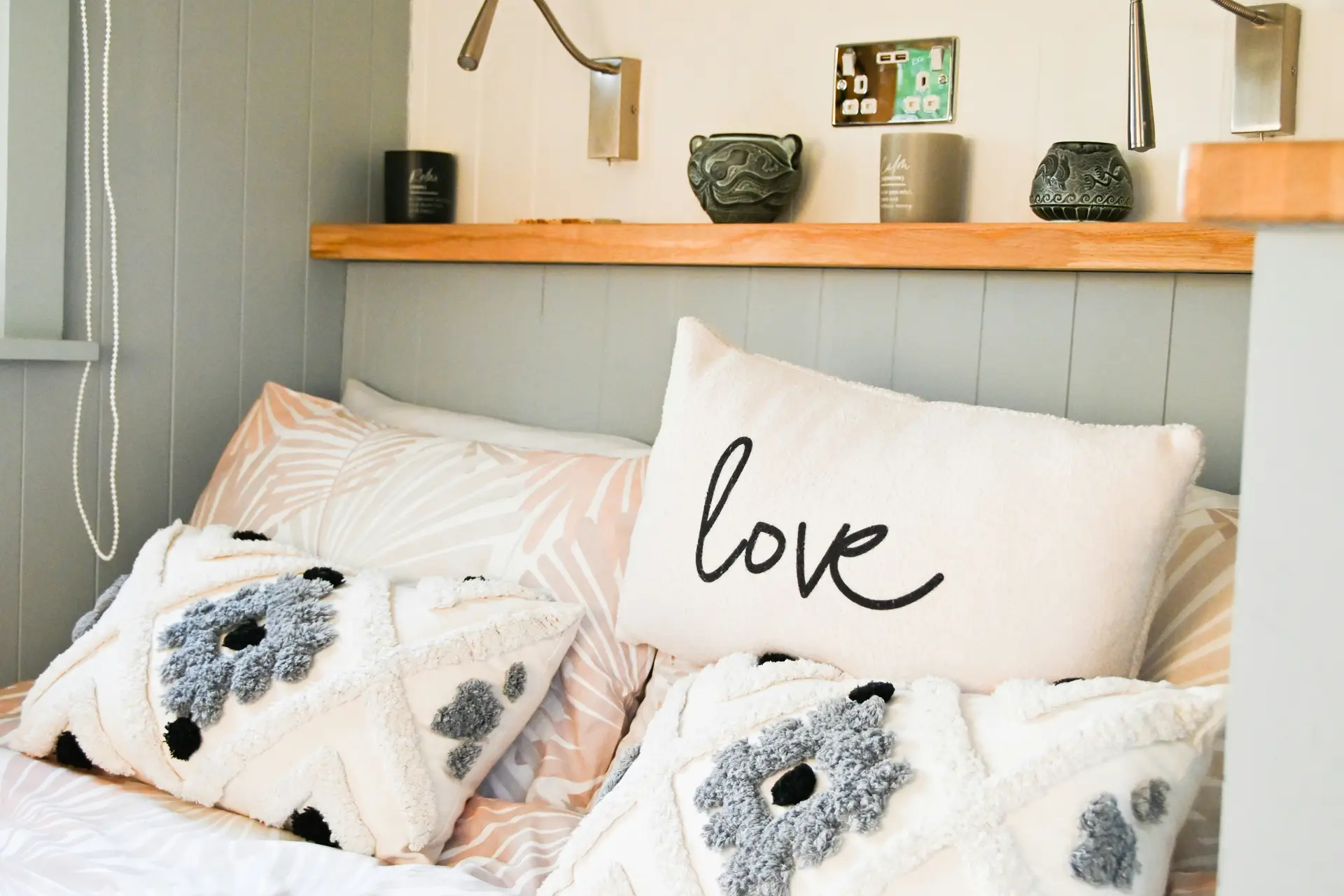 Cozy bed decor with pillows featuring a creative pattern mix plus saying for a playful yet inviting look. Textured cushions with bold tufted designs in gray and white are paired with a soft pillow that simply reads “love,” adding warmth and personality. Subtle printed sheets and a wooden shelf with decorative accents complete the versatile and stylish setup.