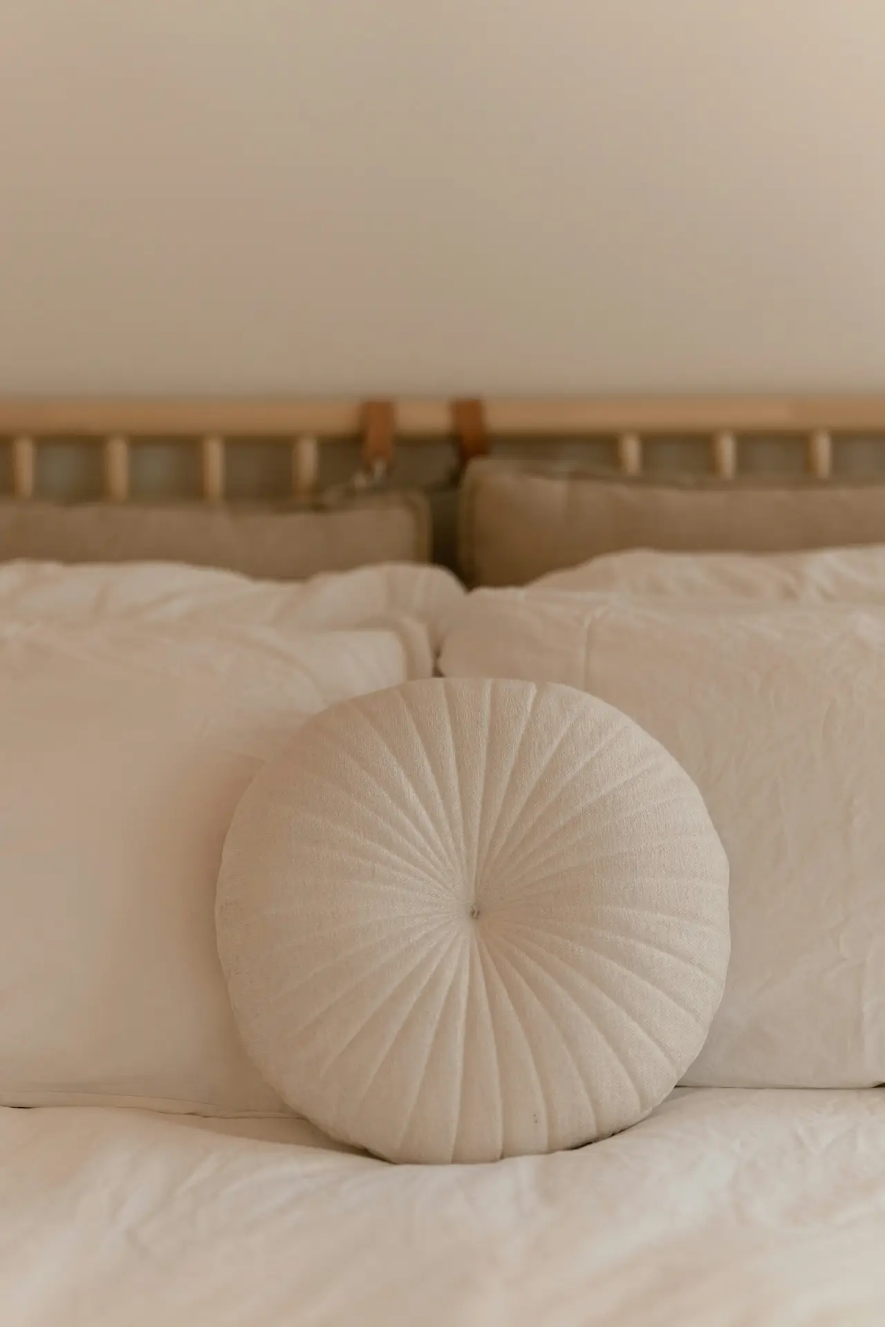 Neutral-toned bed decor with pillows arranged in layers of soft beige and white, creating a calm and cozy look. A standout circular cushion at the center shows how to play with shapes, breaking up the usual rectangular lines of bedding. The simple wooden headboard adds to the natural, versatile aesthetic, making the space feel both modern and timeless.