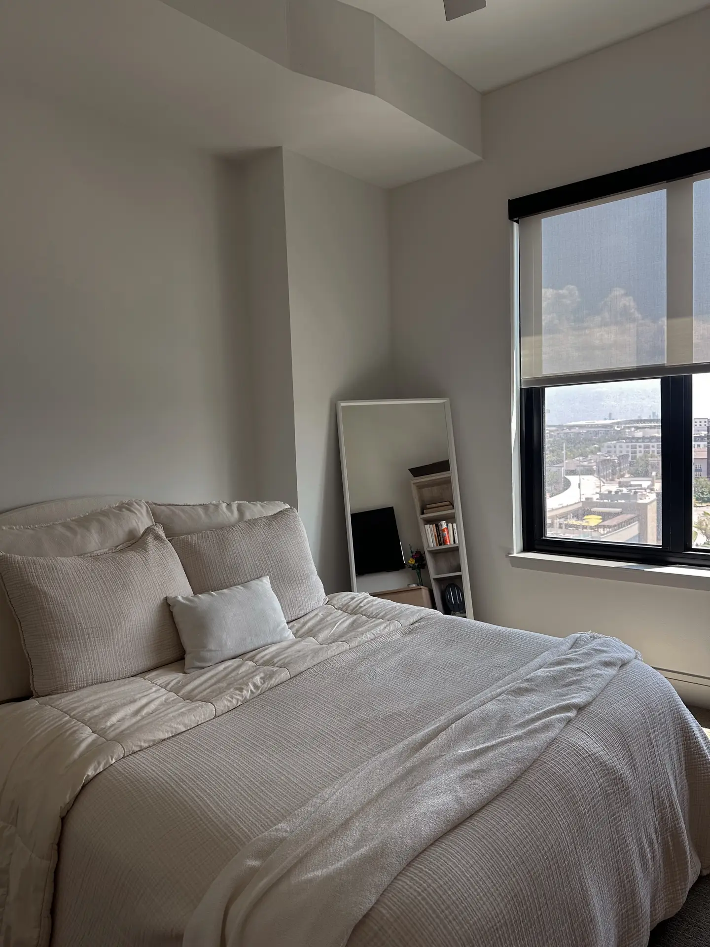 A minimal bedroom styled with layered bed decor with pillows, arranged using the rule of odds for a balanced look. Three large neutral cushions anchor the bed, paired with two smaller accent pillows in soft tones. A tall mirror, books, and a window with city views add a clean and modern touch to the serene space.