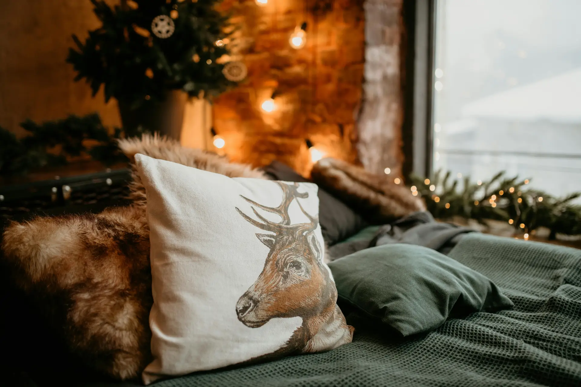 Warm and festive bed decor with pillows styled for a cozy winter setting, featuring a reindeer-print cushion, dark green pillow, and soft faux fur textures. The setup highlights the idea of seasonal swapping, where decorative accents and fabrics can be updated to match the mood of the season. Twinkling string lights, a Christmas tree, and a snowy view outside the window enhance the inviting atmosphere.