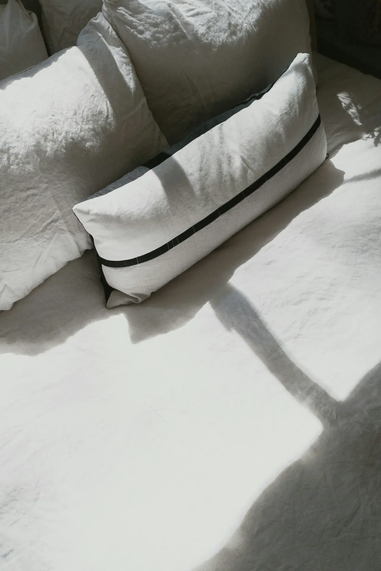 Minimalist bed decor with pillows in soft neutral tones, highlighted by a long rectangular cushion featuring a single black line across its center. The natural light and shadows falling across the bedding create a serene, calming effect. This simple yet refined setup shows how subtle details can elevate a space without overwhelming it.