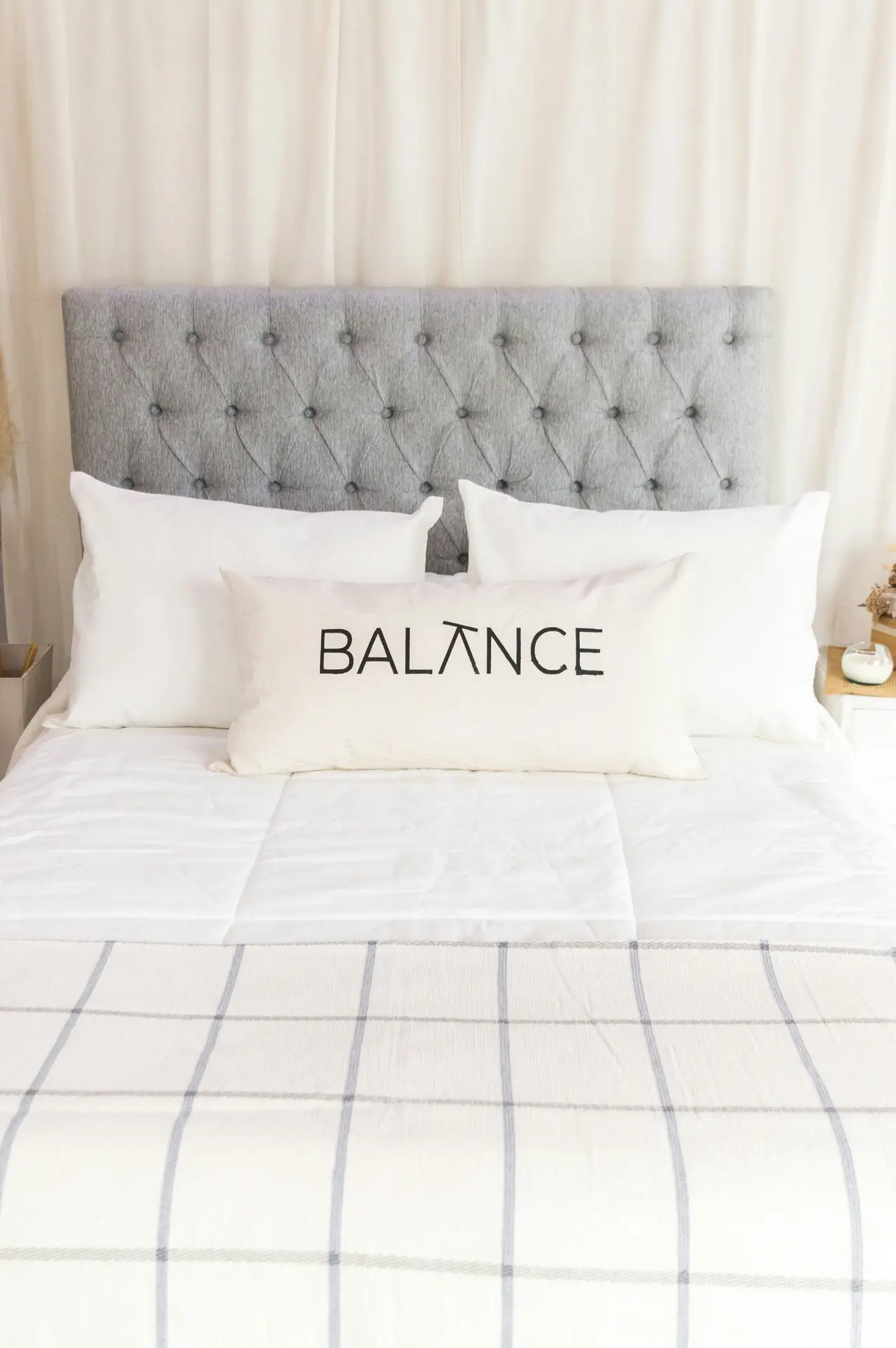 Modern bed decor with pillows styled in crisp white bedding, layered against a tufted gray headboard for a clean and elegant look. A bold statement pillow with the word “BALANCE” in black lettering takes center stage, adding personality and focus to the setup. The checkered throw at the foot of the bed ties the look together, offering a versatile mix of simplicity and style.