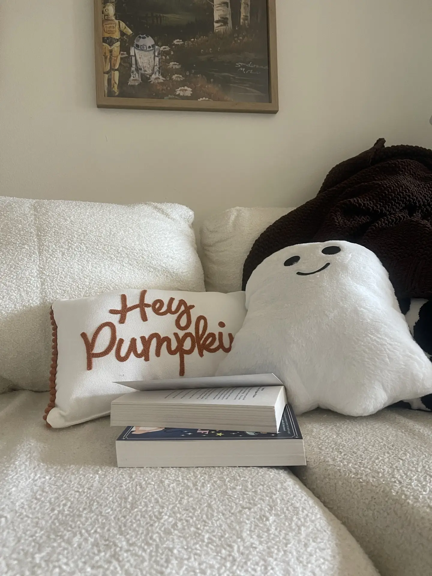 Playful bed decor with pillows featuring cozy white seating accented by themed pillows, including a “Hey Pumpkin” design and a smiling ghost cushion. A dark knit blanket draped in the corner adds warmth, while a couple of books on the bed suggest a relaxed, seasonal reading nook. The quirky wall art above enhances the personalized and inviting atmosphere.