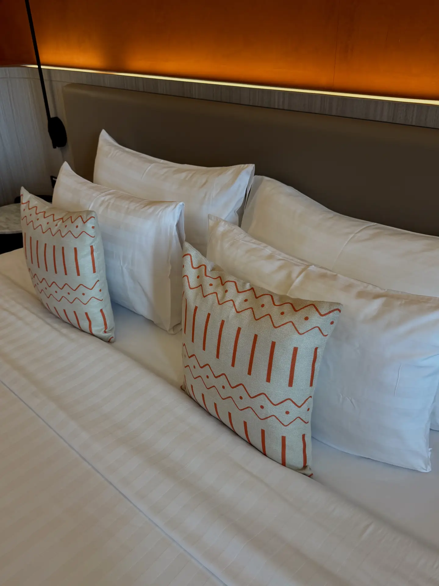 Modern bed decor with pillows featuring crisp white bedding and a mix of plain and patterned cushions. The arrangement shows how to use pillows in different sizes for both comfort and style, with larger white pillows layered at the back and smaller decorative ones in front with orange geometric designs. A warm backlight above the headboard adds a cozy yet contemporary touch, making the setup versatile for various bedroom styles.