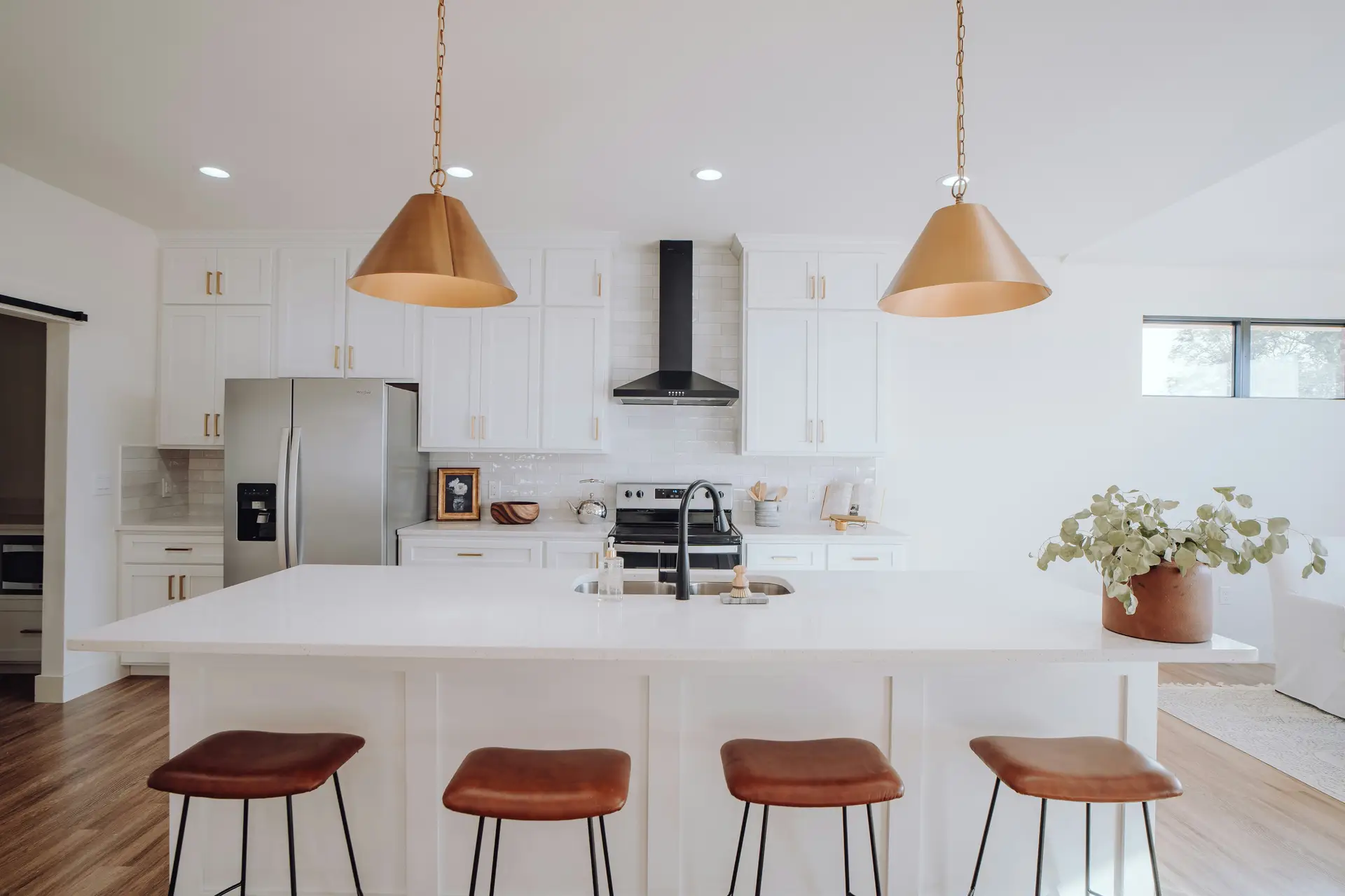 Bright white kitchen with shaker cabinets, stainless steel appliances, and a spacious island with leather barstools. Two oversized brass cone pendant lights hang above, adding warmth and elegance while highlighting modern kitchen lighting ideas that combine functionality with stylish metallic accents.