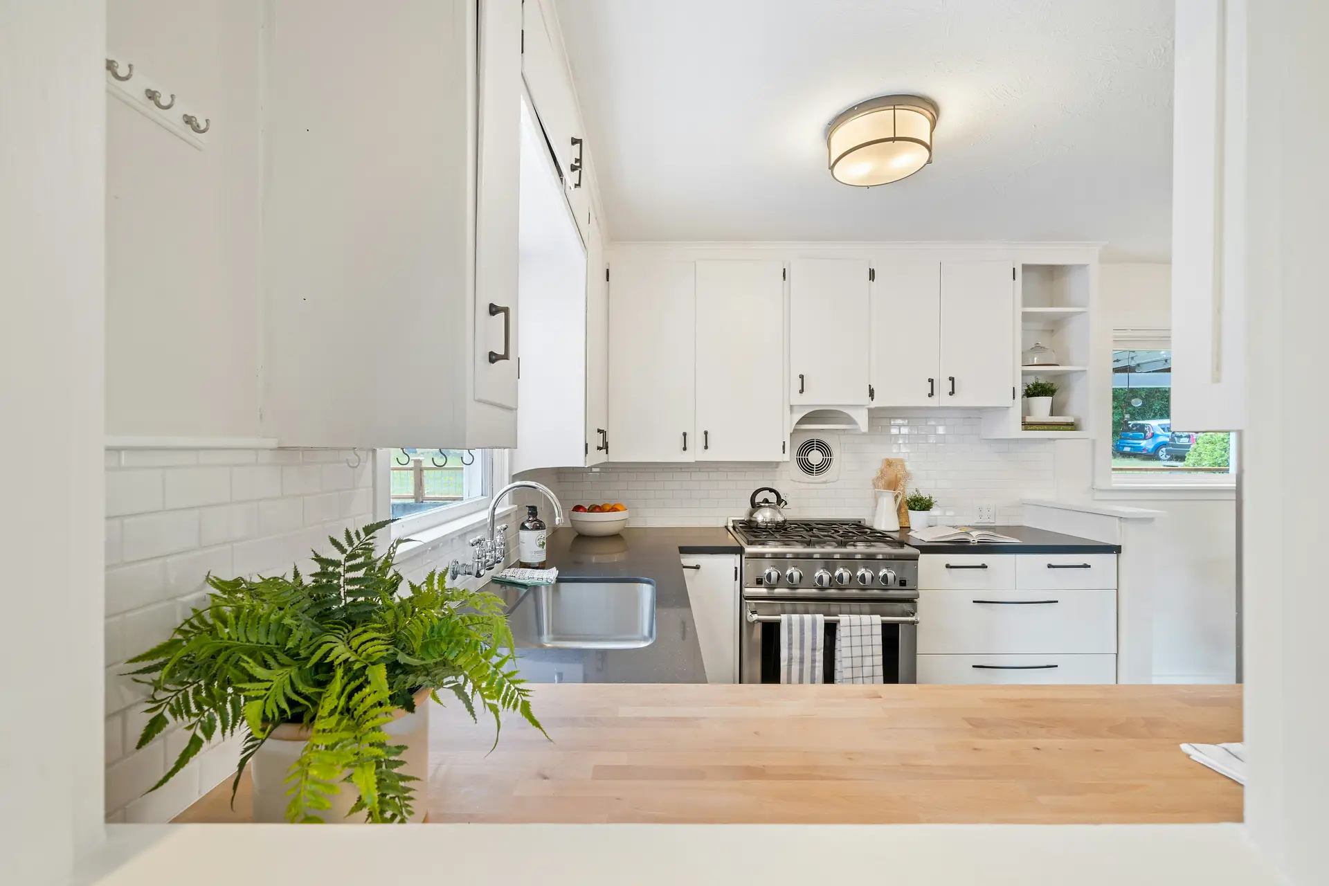 Bright white kitchen with shaker-style cabinets, subway tile backsplash, and stainless steel appliances, illuminated by a flush mount ceiling light. A wooden countertop, greenery, and minimal décor highlight clean design while offering practical kitchen lighting ideas that work in both modern and traditional homes.