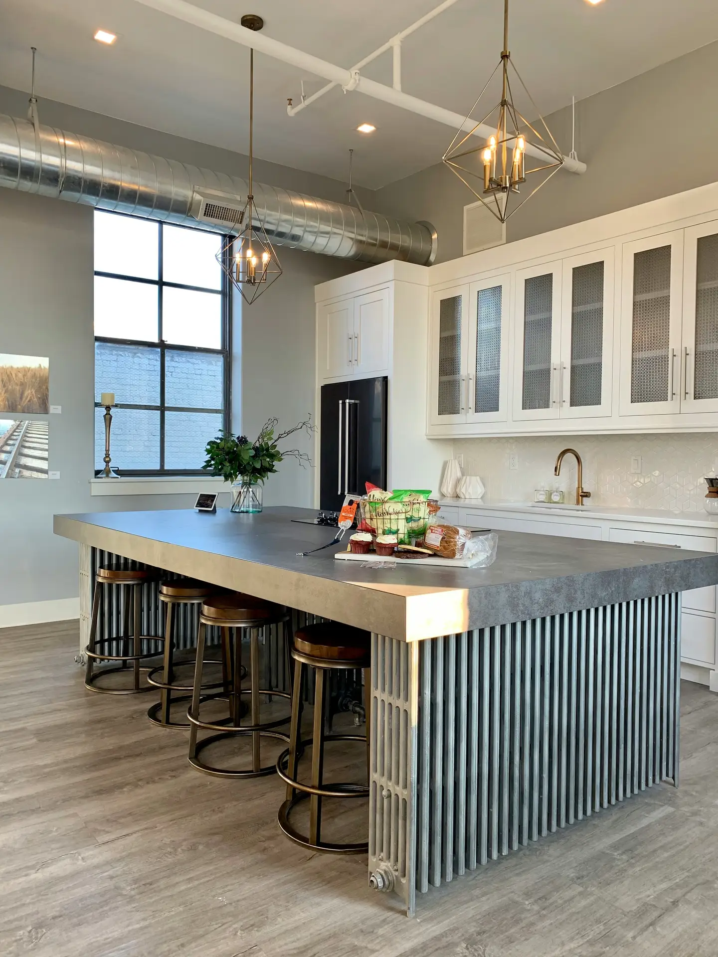 Industrial-style kitchen featuring a large concrete island with metal base, paired with backless stools and white cabinetry. Overhead, striking geometric pendant lights bring a modern design statement, showcasing creative kitchen lighting ideas that blend functionality with bold architectural style.