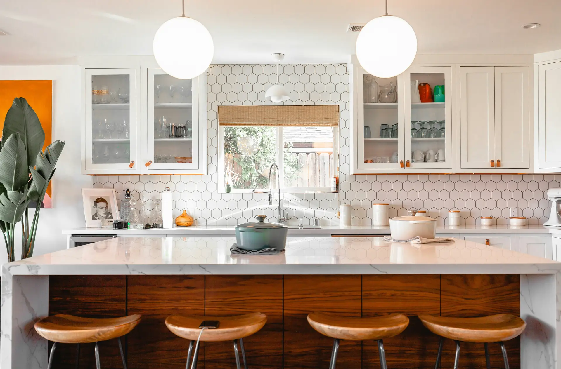 Contemporary kitchen with white hexagon tile backsplash, glass-front cabinets, and a large marble island paired with wooden stools. Overhead, two round globe pendant lights provide a sleek focal point, serving as stylish kitchen lighting ideas that balance modern design with warm functionality.