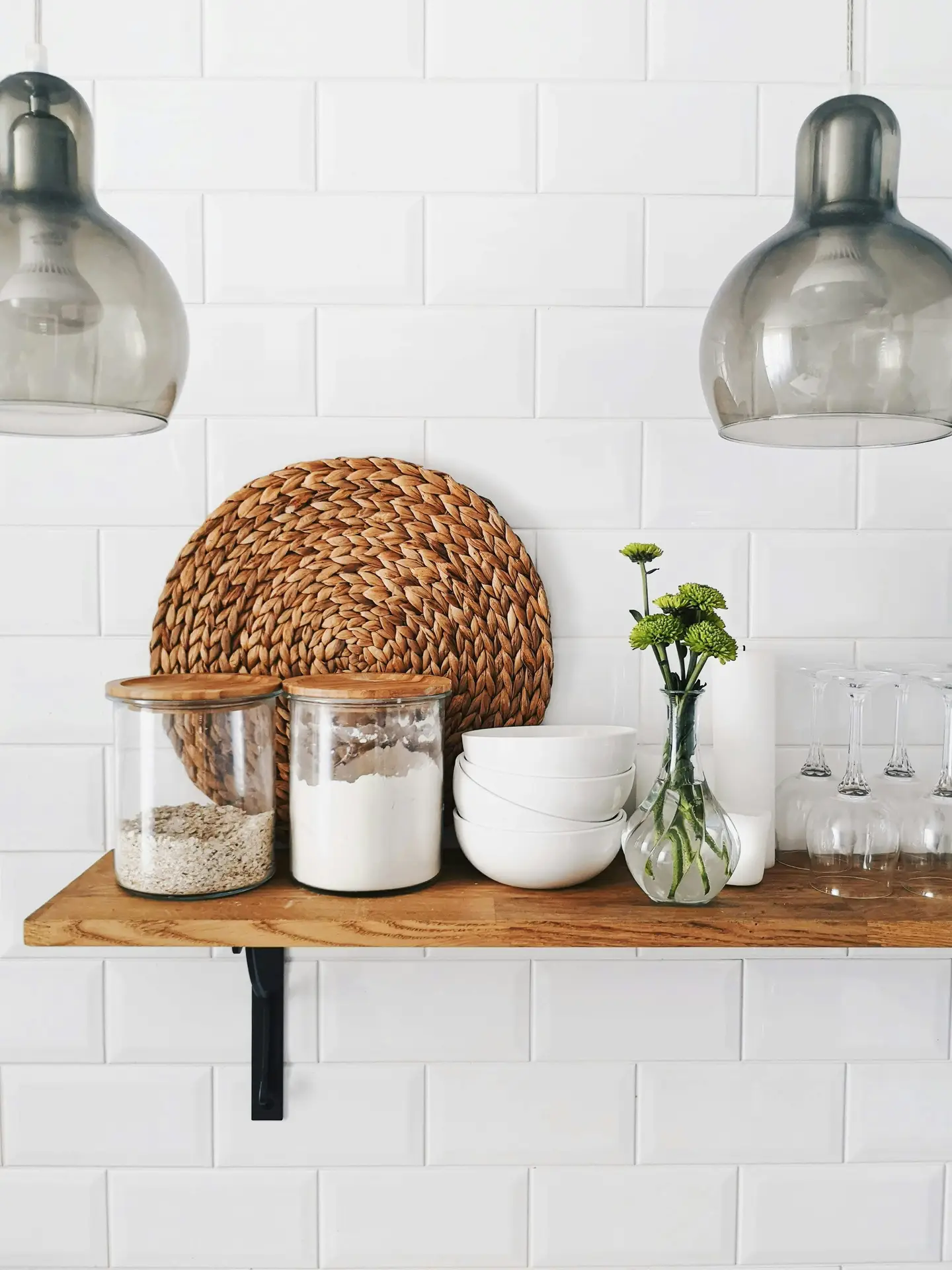 Minimalist kitchen setup with white subway tile backsplash and a wooden floating shelf styled with glass jars, white bowls, woven décor, and a small vase of flowers. Two sleek hanging lights above floating shelf add contrast and ambiance, making this a great example of modern kitchen lighting ideas that balance function with stylish simplicity.