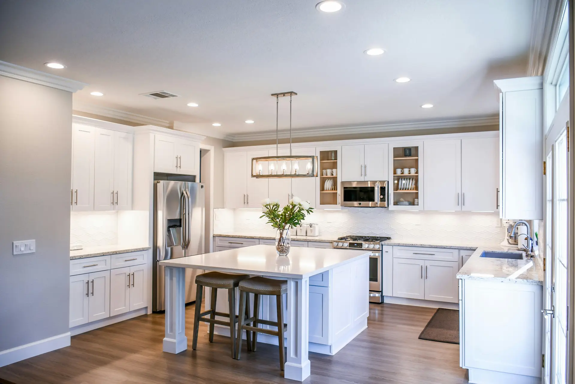 Spacious white kitchen with stainless steel appliances, hardwood flooring, and a central island decorated with fresh flowers. A rectangular fixture with multiple bulbs serves as elegant hanging lights above kitchen island, complementing recessed ceiling spots and offering versatile kitchen lighting ideas for both task and ambient illumination.