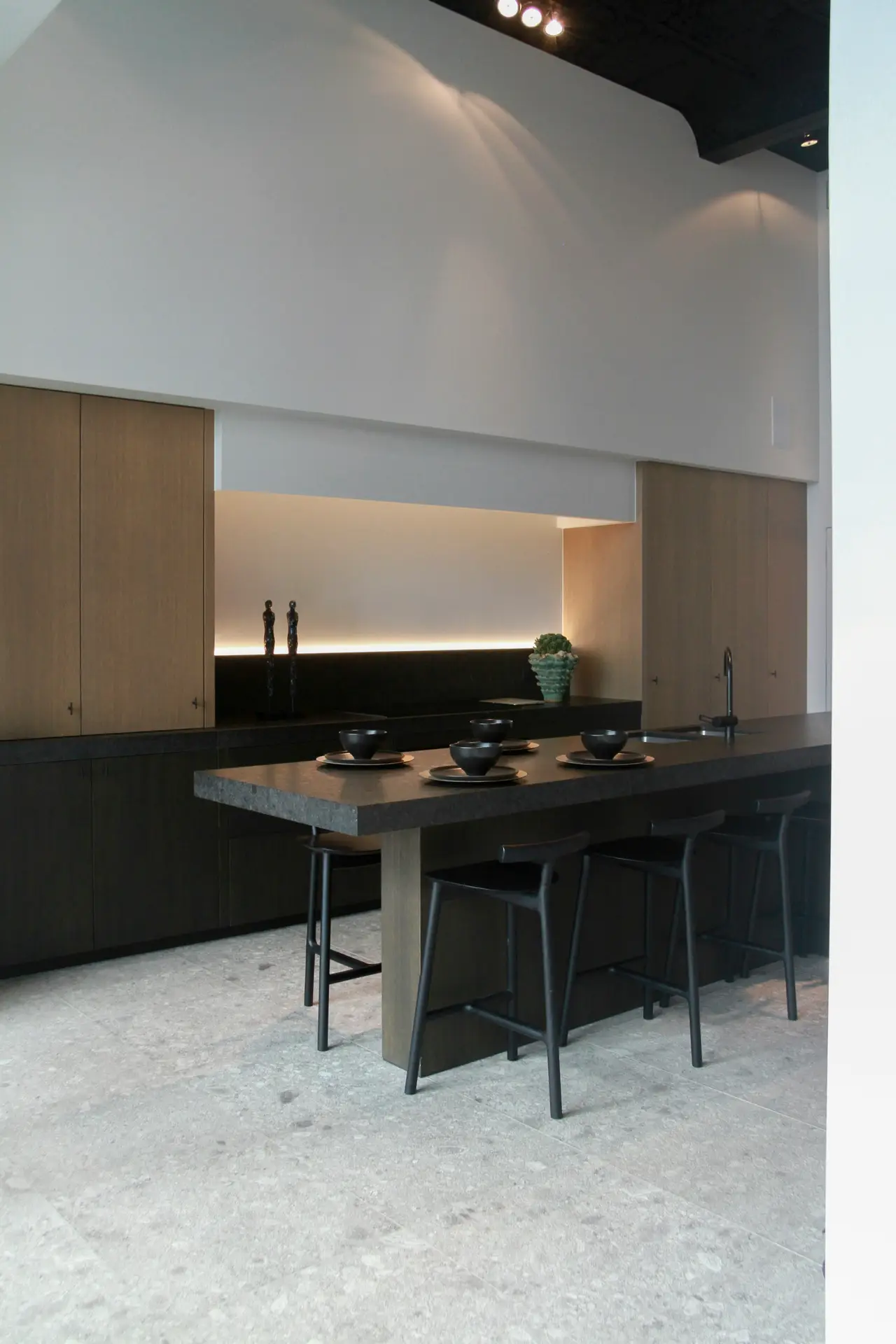 Modern minimalist kitchen with dark cabinetry, a sleek stone island, and black barstools arranged for dining. Subtle LED strip lighting runs along the backsplash, adding soft ambient glow and highlighting clean lines. An example of kitchen lighting ideas that combine elegance, warmth, and functionality.