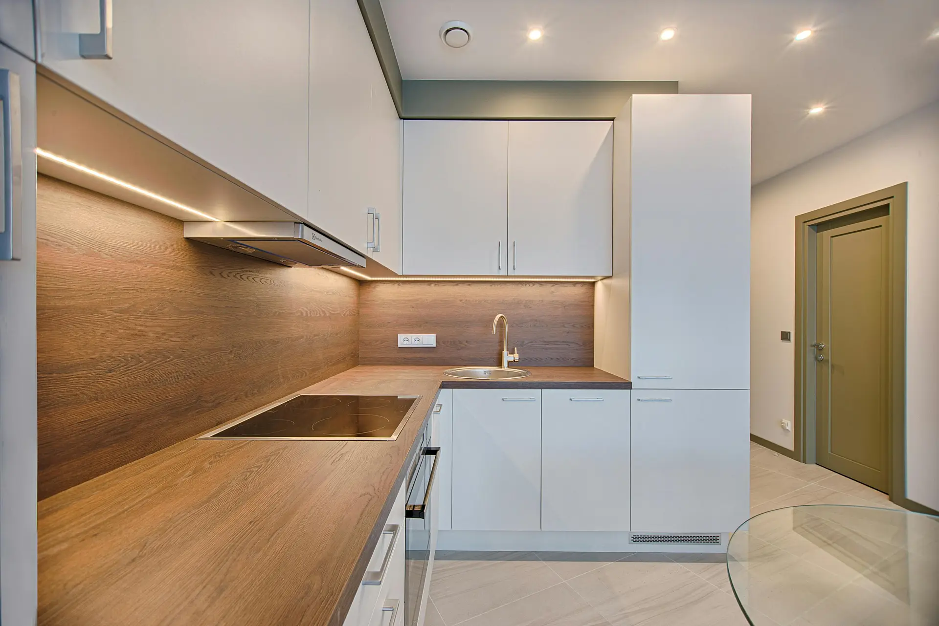 Modern L-shaped kitchen with wood countertops, white cabinets, and a sleek induction cooktop. A clever lighting mix of recessed ceiling spots and under-cabinet LEDs brightens the workspace, providing practical kitchen lighting ideas that balance task illumination with a warm, inviting atmosphere.