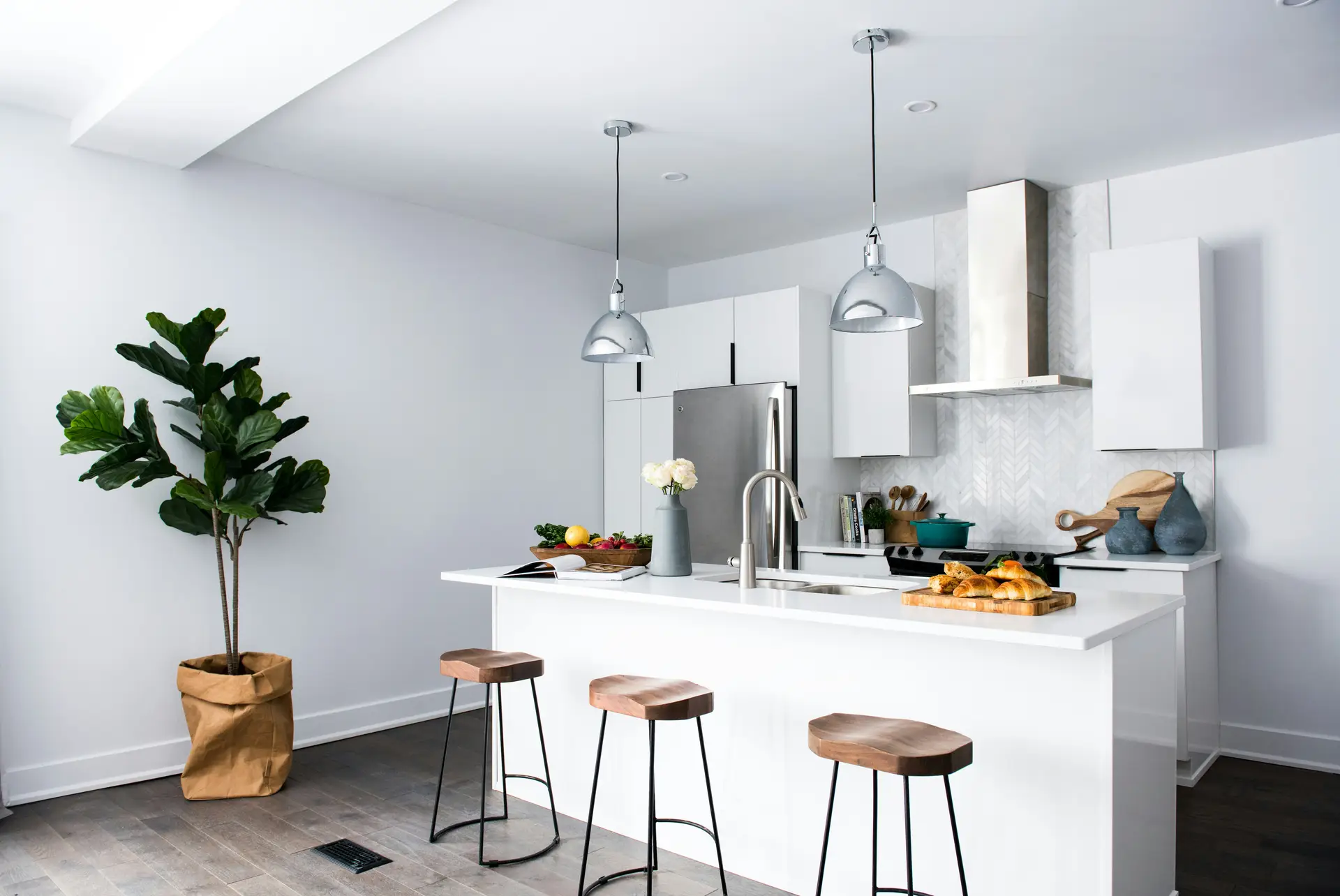 Stylish kitchen with white cabinetry, stainless steel appliances, and a central island paired with wooden stools. Overhead, sleek metal pendant lights provide focused illumination, highlighting fresh produce and baked goods while serving as functional kitchen lighting ideas that suit modern and contemporary interiors.