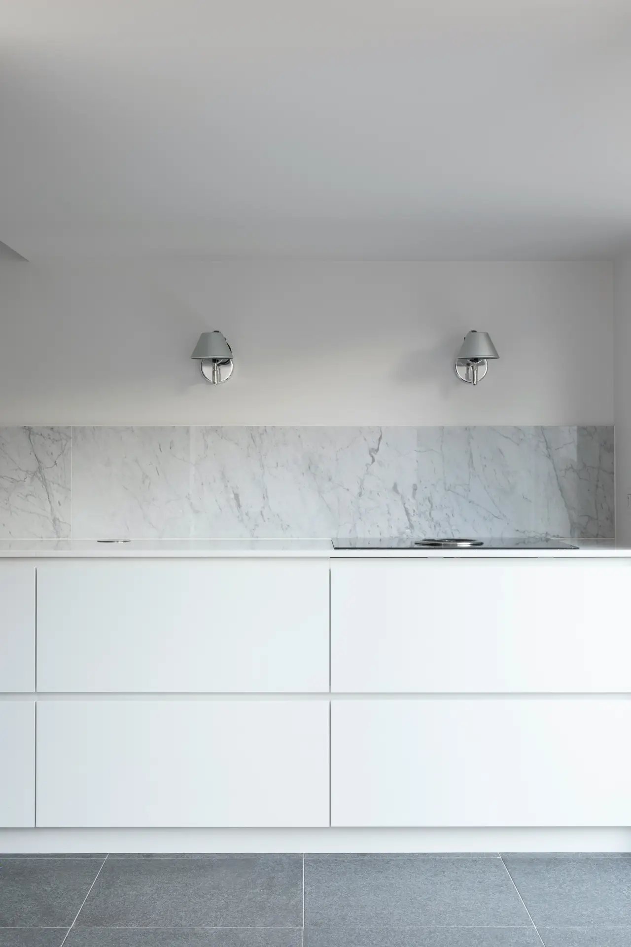 Minimalist white kitchen with sleek cabinetry, marble backsplash, and clean gray tile flooring. Two modern metal wall sconces add a polished touch while providing task lighting, showcasing kitchen lighting ideas that enhance a streamlined and contemporary design.