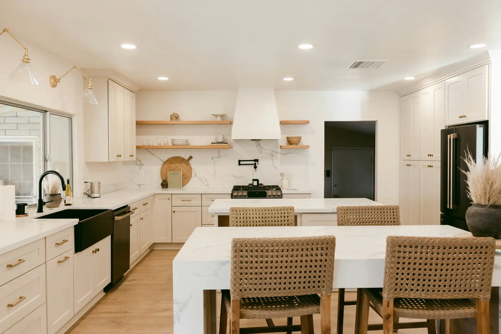 Elegant open kitchen with white cabinetry, marble-look counters, and woven barstools creating a warm modern feel. Multiple recessed ceiling lights provide bright, even illumination, paired with glass sconces above the sink, offering layered kitchen lighting ideas that combine functionality with stylish design.