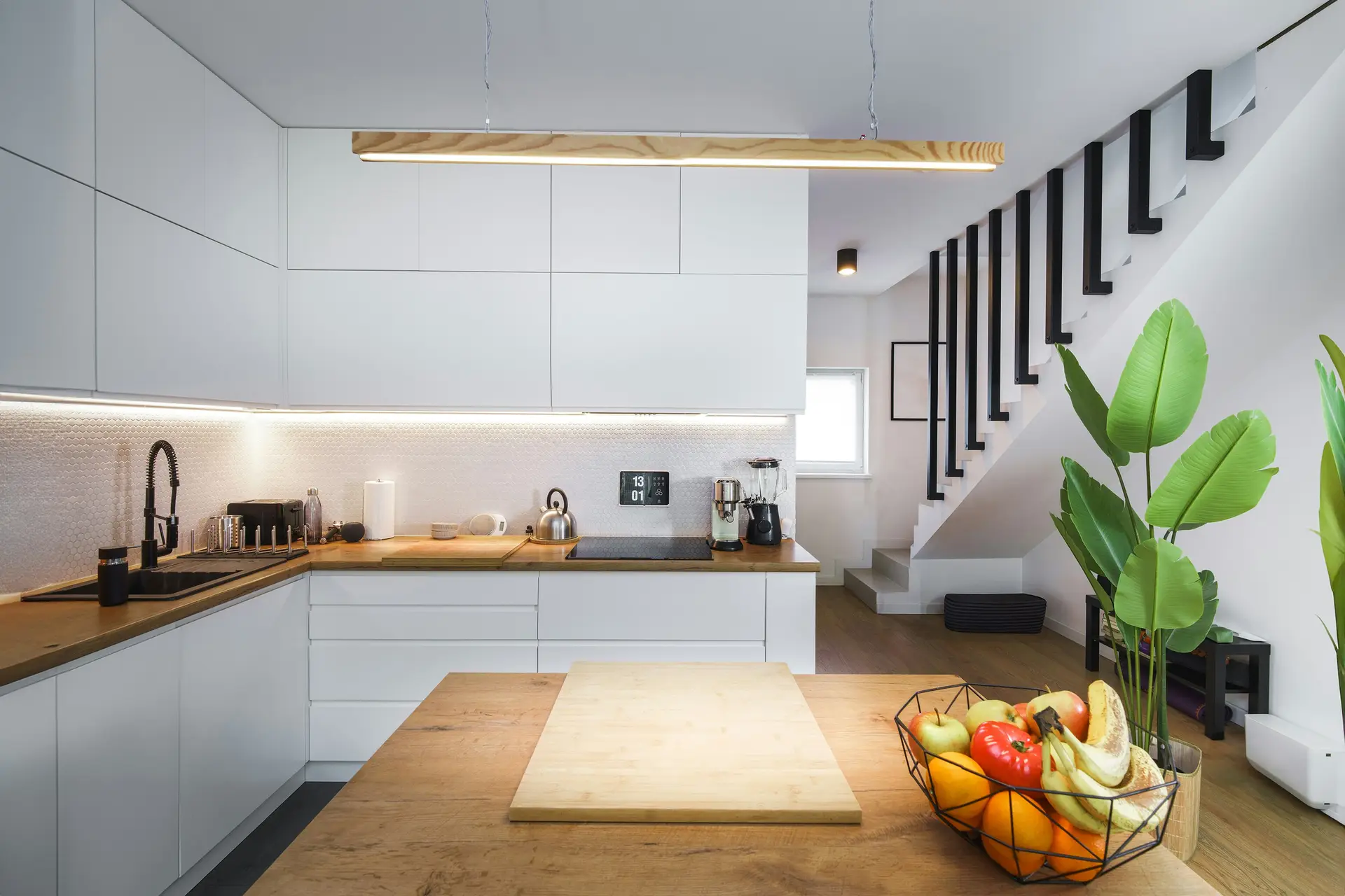 Contemporary open kitchen with sleek white cabinets, wood countertops, and a central island decorated with fresh fruit and greenery. A minimalist wooden linear pendant light hangs above, paired with subtle under-cabinet illumination, offering modern kitchen lighting ideas that merge natural warmth with clean, functional design.