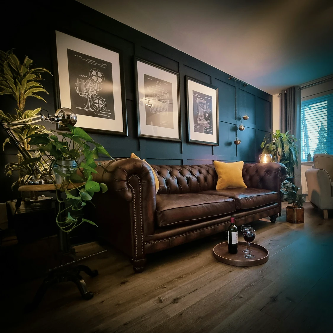 Stylish living room featuring a deep blue accent wall with three framed prints as focal points, paired with a tufted leather Chesterfield sofa and mustard throw pillows. Lush greenery, warm lighting, and rustic wood flooring complete the cozy yet elegant atmosphere, offering inspiring living room decor ideas for walls that mix art, texture, and bold color.