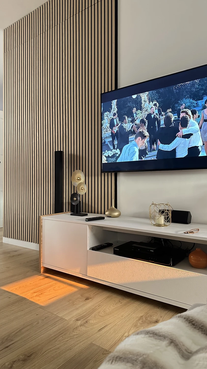 Modern living room with sleek vertical wall paneling in natural wood tones, creating a stylish backdrop for the mounted TV. A minimal white media console holds decorative accents in gold and black, blending function with elegance. This setup highlights smart living room decor ideas for walls, showing how texture and contrast can elevate a cozy entertainment space.