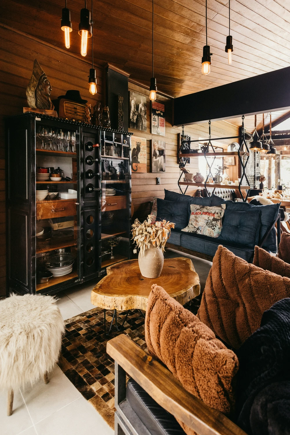 Rustic farmhouse-inspired living room featuring a raw-edge wooden coffee table as the centerpiece, paired with cozy textured pillows, a fur-covered stool, and warm pendant lighting. The space blends vintage accents, open shelving with glass doors, and layered wall art, offering creative living room farmhouse decor ideas that combine comfort with character.