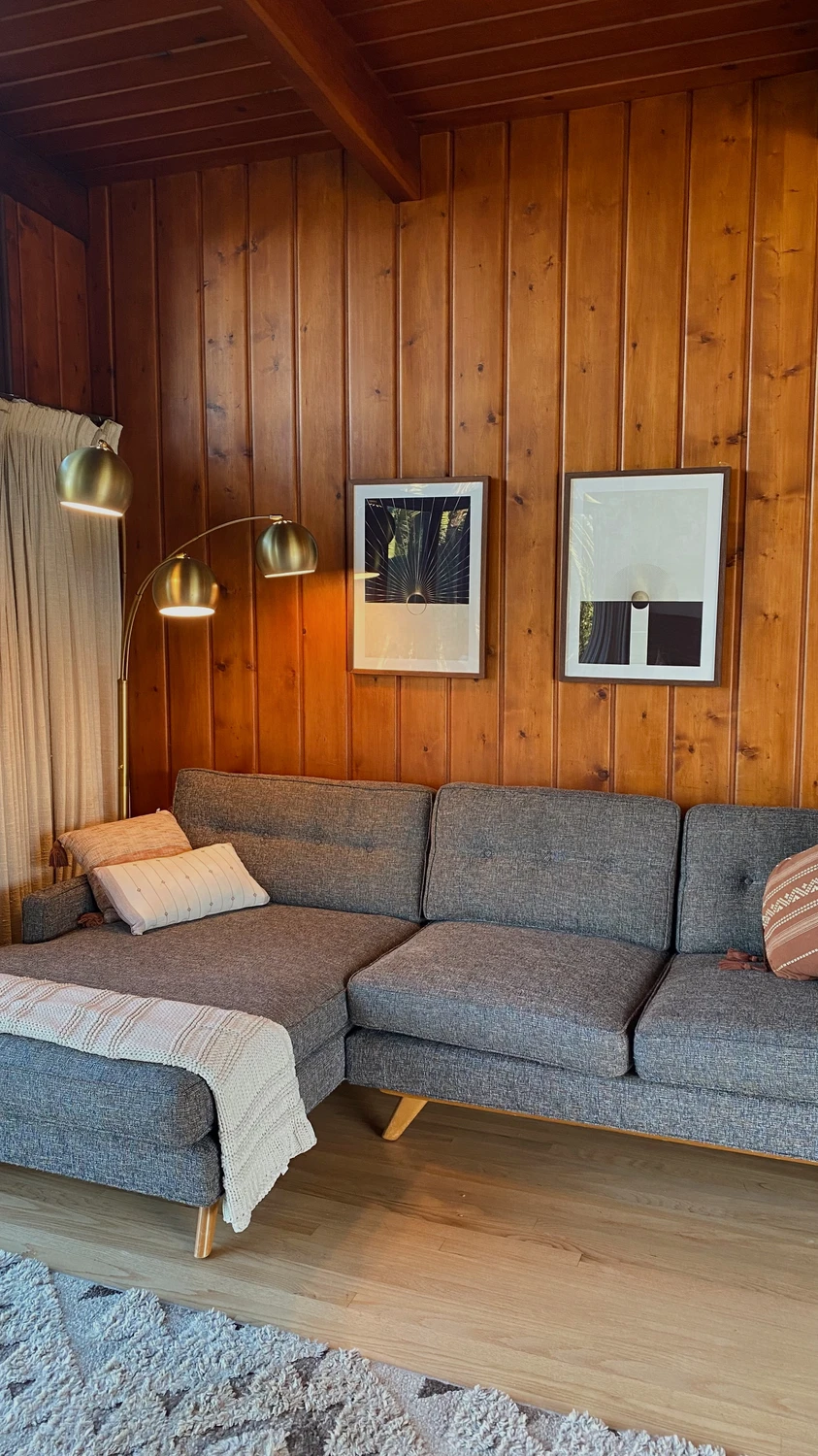Living room with a cozy l-sectional sofa in gray fabric, styled with neutral throw pillows and a knitted blanket, perfect for modern living room layout ideas. The wooden paneled wall adds warmth, decorated with abstract framed artwork and a brass floor lamp with multiple lights, creating a balance of mid-century charm and functional comfort.