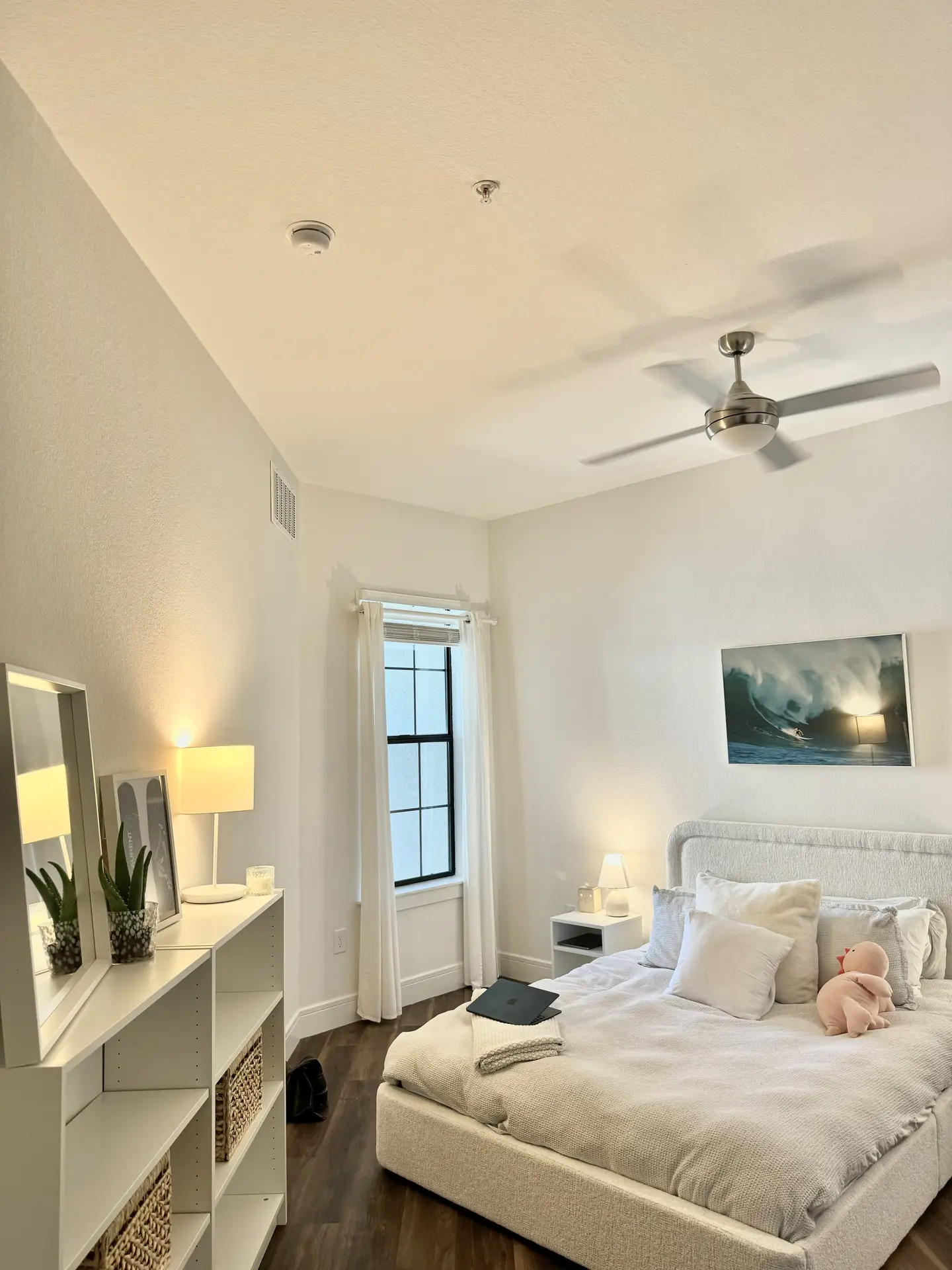 Bright and airy bedroom styled with an all white palette, using a crisp paint color for the bedroom that enhances natural light. The soft upholstered bed, layered with neutral bedding and pillows, creates a cozy yet minimalist feel. Simple furniture, warm lighting, and a wave-inspired wall art piece complete the fresh and versatile design.