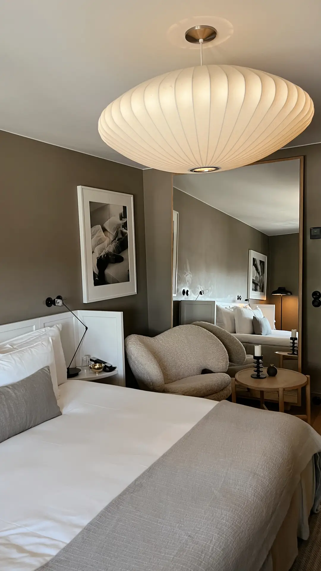 Modern bedroom styled in a warm paint color for the bedroom that leans toward elegant beige, creating a calm and sophisticated backdrop. The bed is dressed in crisp white linens with gray accents, complemented by a cozy armchair and minimalist side tables. Large mirrors, framed black-and-white photography, and a sculptural ceiling lamp enhance the versatile, contemporary atmosphere.