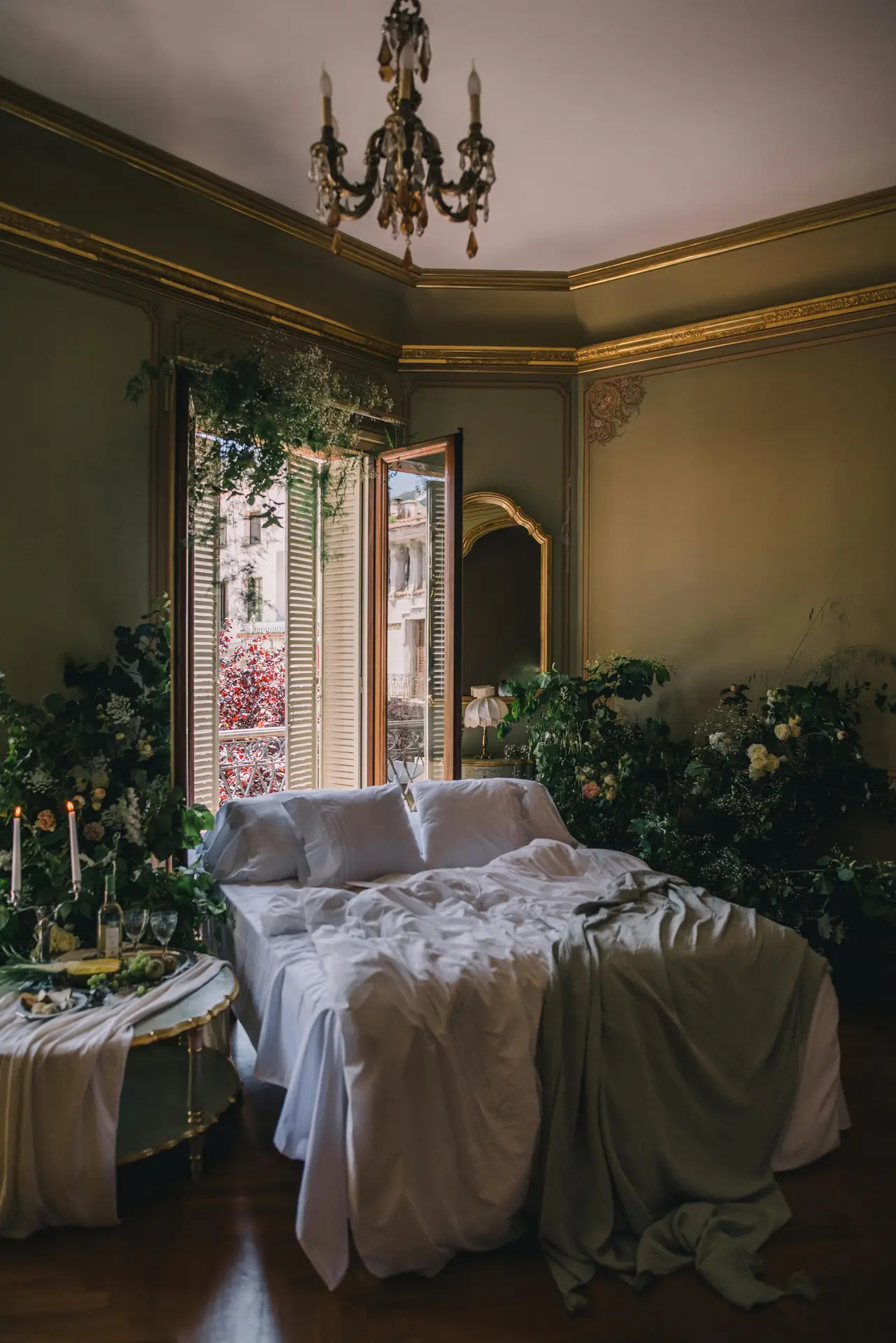 Elegant bedroom styled with a paint color for the bedroom in botanical green with gold accents, creating a luxurious and timeless atmosphere. The ornate molding, chandelier, and antique mirror complement the rich tones, while lush greenery and flowers bring nature indoors. A cozy bed with crisp white linens and a draped green throw adds softness to the sophisticated setting.