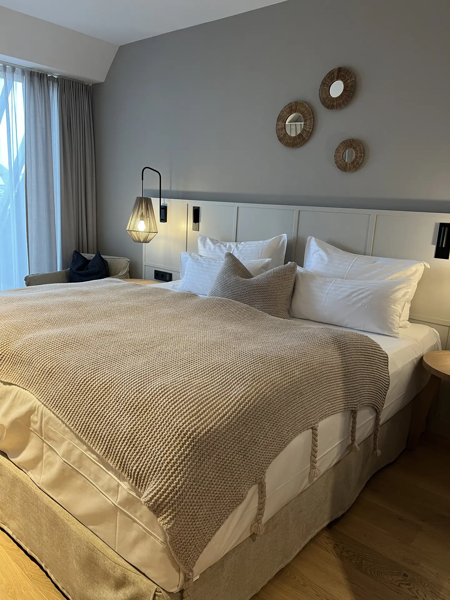 Inviting bedroom designed with a soft paint color for the bedroom in cozy gray, creating a soothing and modern backdrop. The bed is layered with crisp white pillows, neutral-toned cushions, and a textured knit blanket that enhances warmth. Pendant lighting, natural wood accents, and small decorative mirrors on the wall complete the comfortable yet stylish look.