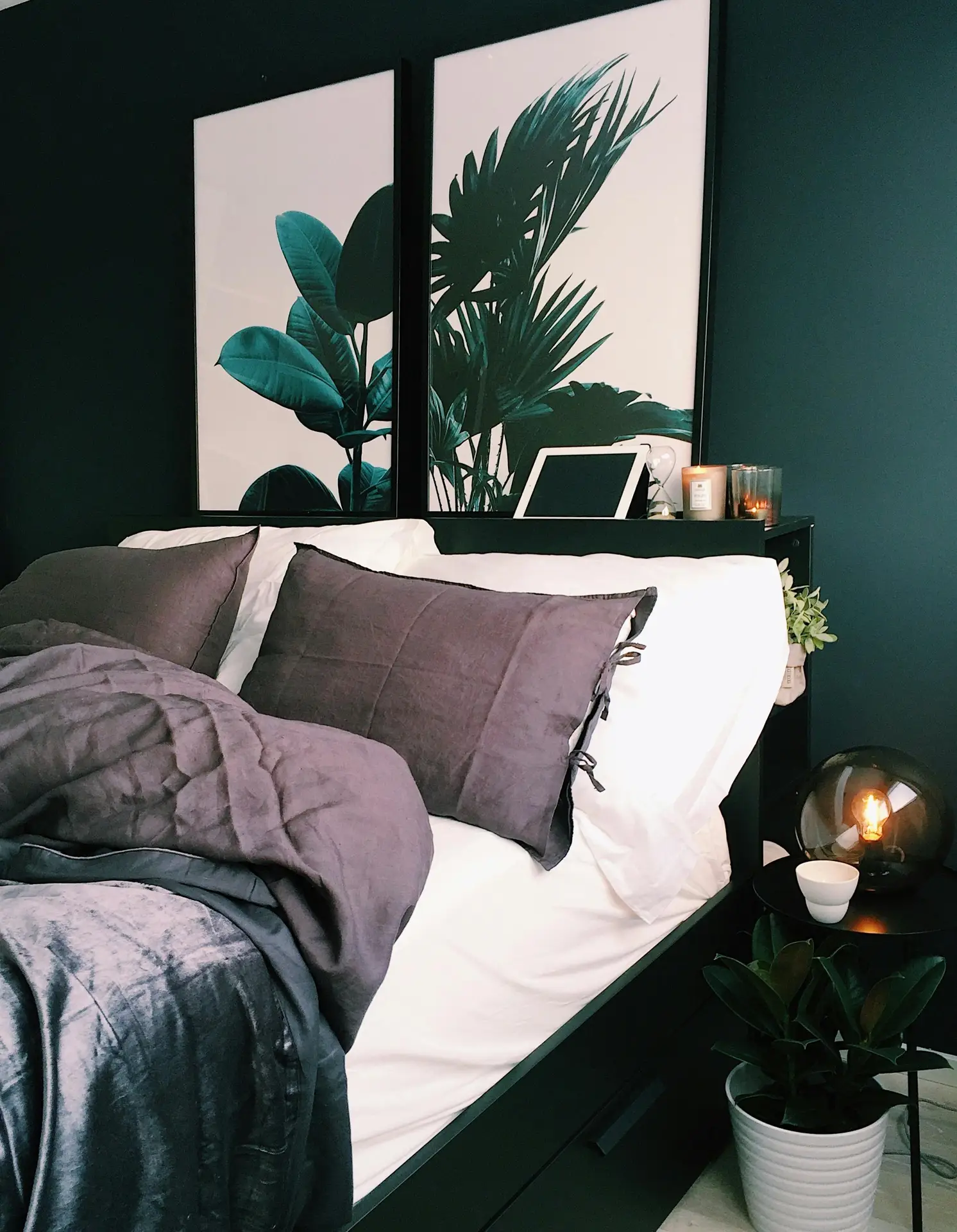 Stylish bedroom showcasing a bold paint color for the bedroom in rich dark green, creating a dramatic and calming atmosphere. The bed is dressed in crisp white sheets with deep gray bedding and pillows, accented by lush botanical artwork above the headboard. A potted plant, glowing lamp, and candles add warmth and texture, making the space feel both modern and inviting.
