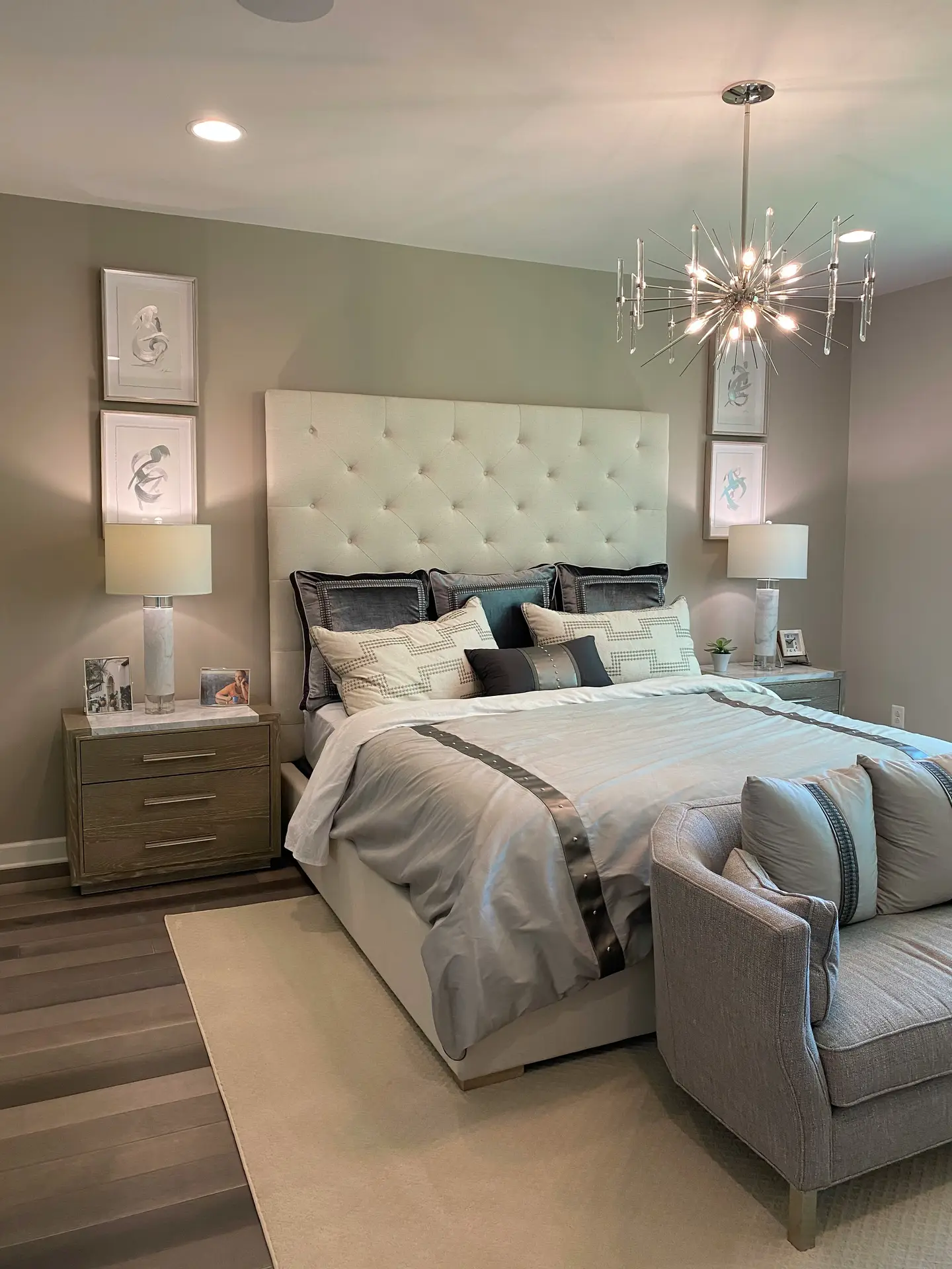 Elegant bedroom designed with a soft paint color for the bedroom in versatile greige, creating a sophisticated backdrop. The tufted headboard, layered bedding in gray tones, and accent pillows give the bed a luxurious feel, while a modern chandelier adds a touch of glamour. Coordinated nightstands, framed artwork, and a cozy seating area complete the balanced and stylish space.