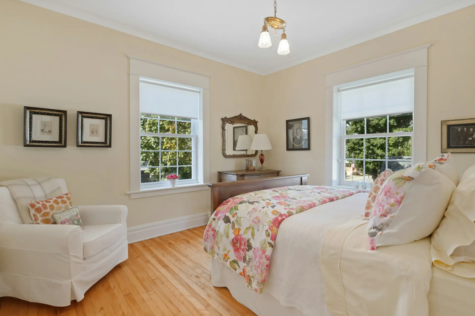 Bright and traditional bedroom styled with a soft paint color for the bedroom in light beige, giving the space a warm and welcoming feel. The bed is dressed in white linens with floral pillows and a matching quilt, adding cheerful pops of color. Hardwood floors, classic furniture, and large windows with natural light complete the cozy, timeless atmosphere.