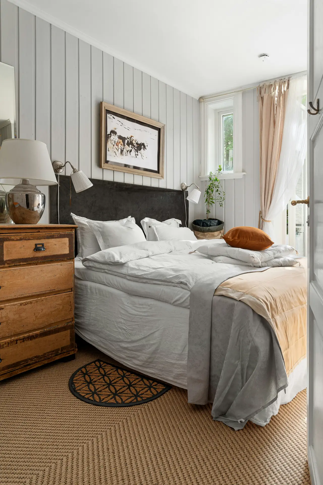 Cozy bedroom styled with paint color for the bedroom in light grey, featuring vertical wood panel walls that add texture and calmness. A rustic wooden dresser contrasts with crisp white bedding, while a soft beige throw and burnt orange cushion bring warmth to the palette. Natural light filters through sheer curtains, highlighting the inviting and balanced design.